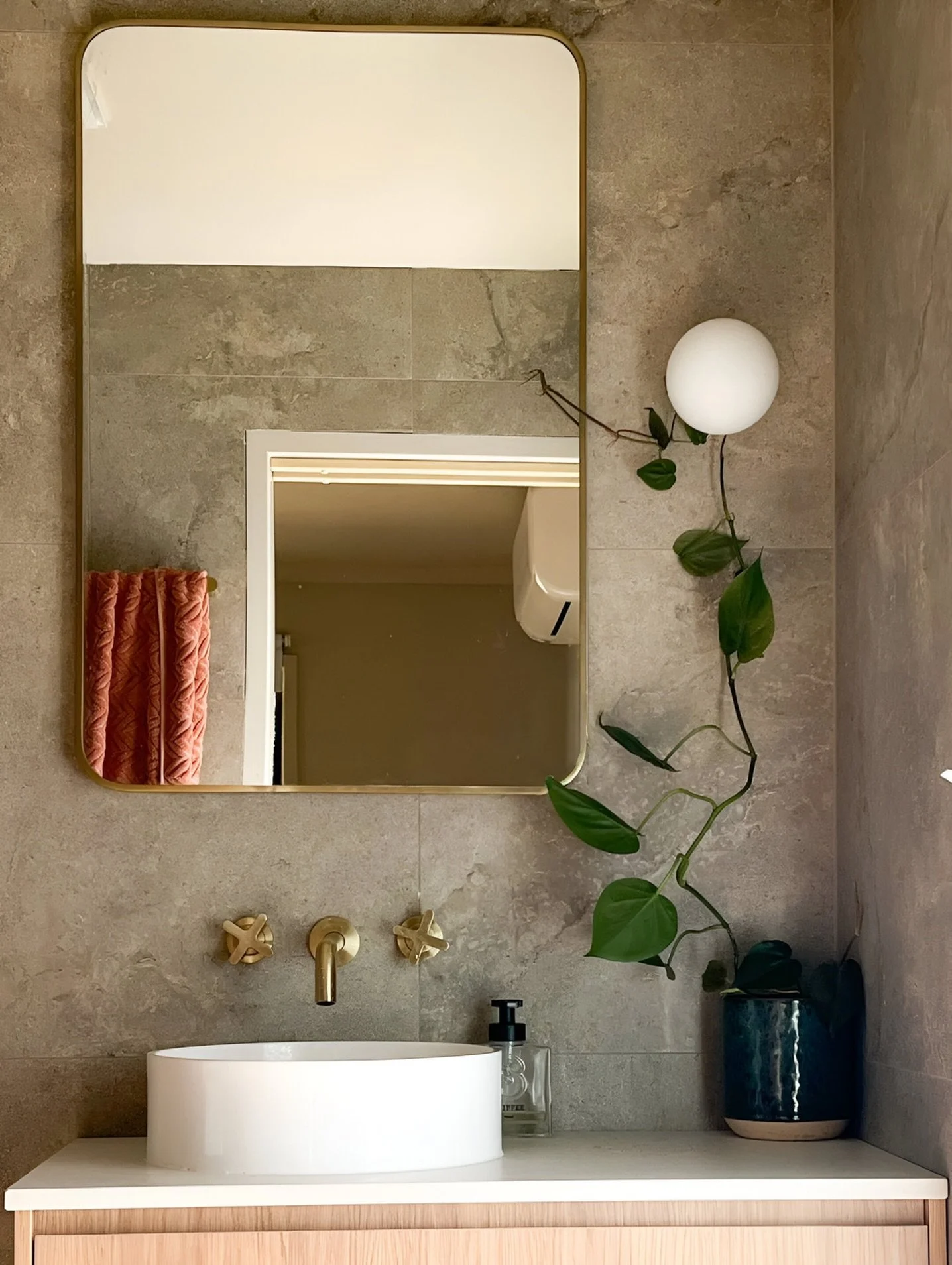 A modern bathroom with a gray tiled wall, a large rectangular mirror, a white vessel sink on a wooden vanity, brass-colored faucet and knobs, a soap dispenser, a potted plant with green leaves, and a spherical wall-mounted light.