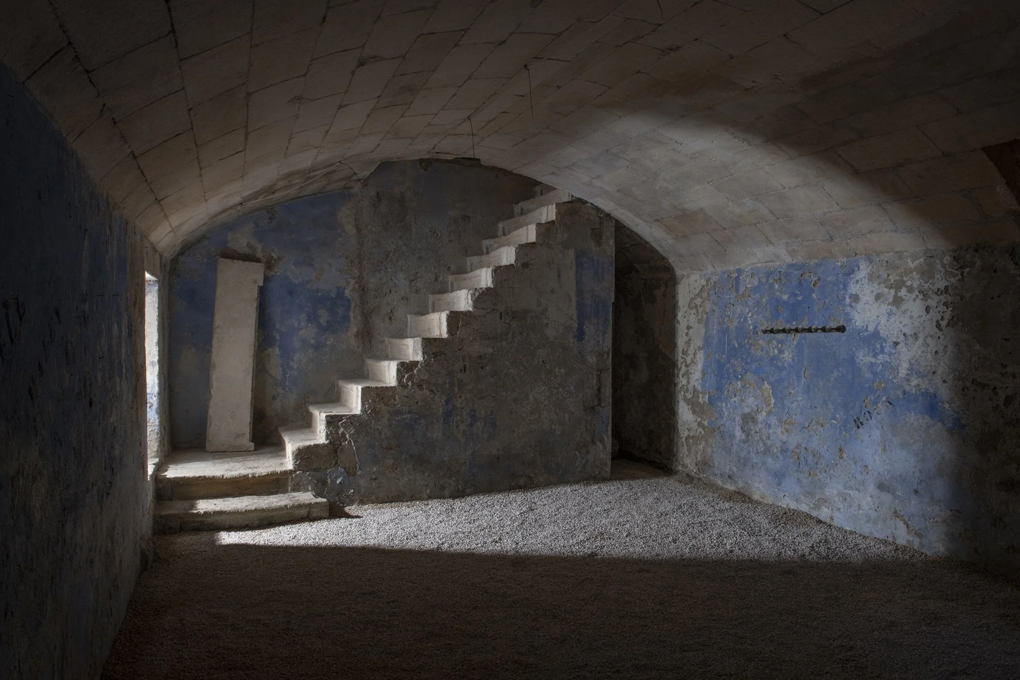 Intérieur d'une pièce ancienne avec murs en pierre, escalier en bois, plafond voûté en pierre, et quelques trouées laissent passer la lumière naturelle.