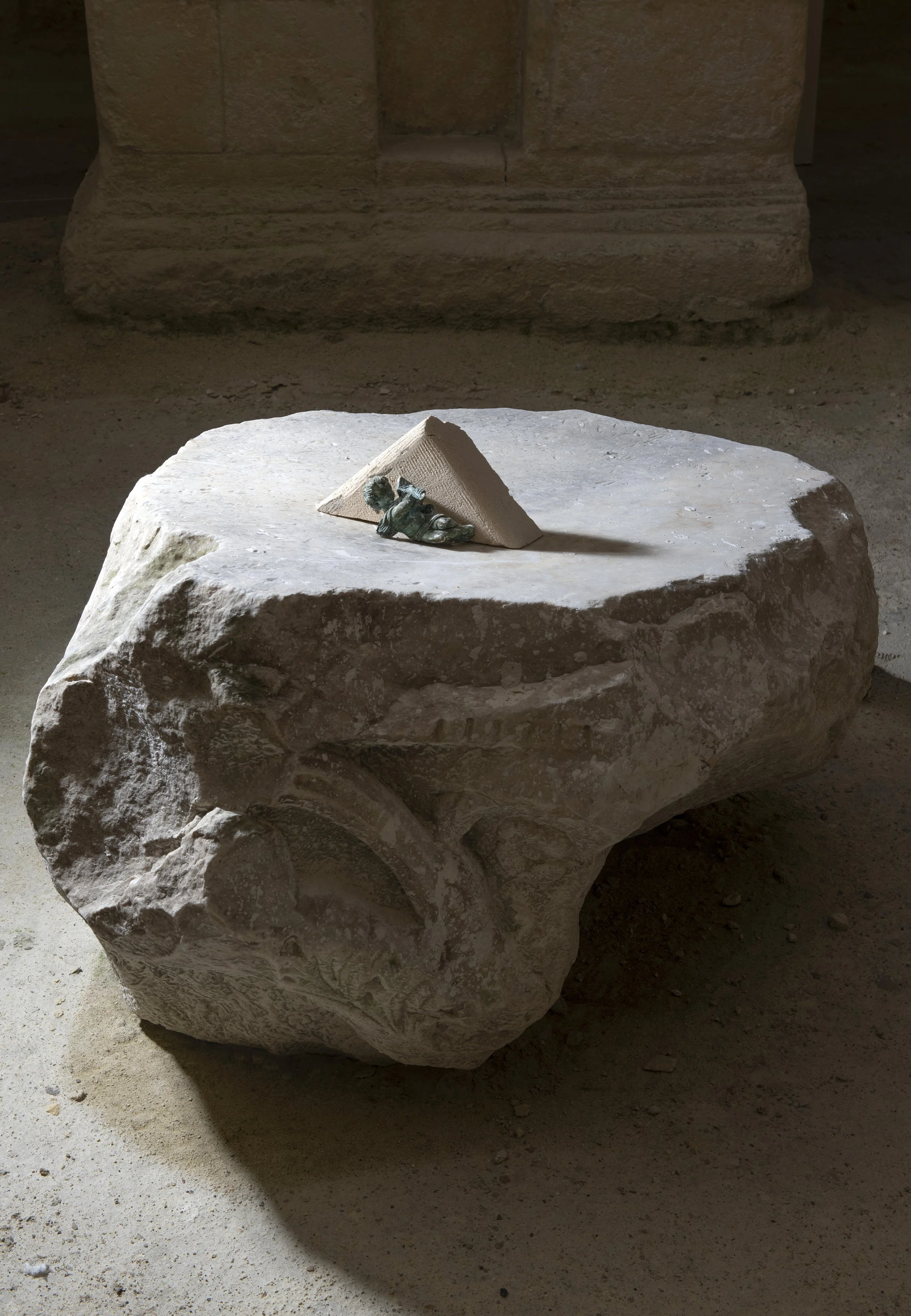 Pierre ancienne avec un morceau de sculpture en bronze, placée dans un espace avec un fond en pierre et sol en terre.