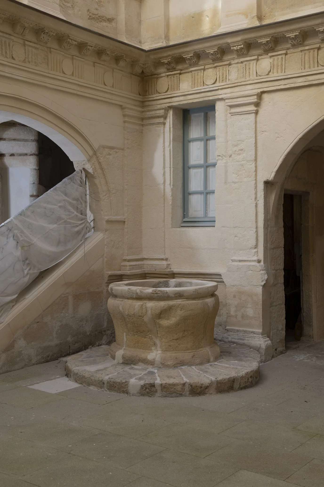Intérieur d'une vieille construction en pierre en cours de restauration, avec une fenêtre en bois peint bleu, un ancien puits en pierre et une arche.