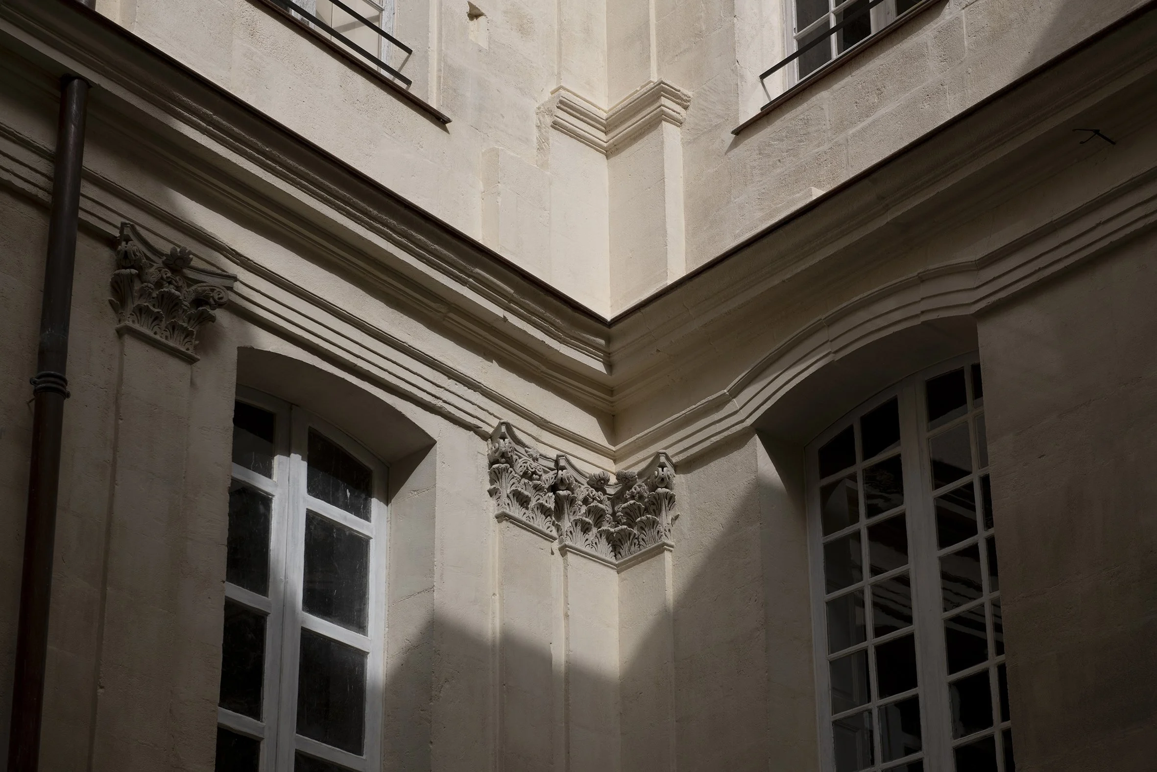 Vue d'un bâtiment avec des fenêtres classiques et des détails architecturaux ornementaux au niveau des coins, avec une lumière naturelle créant des ombres sur la façade en pierre.