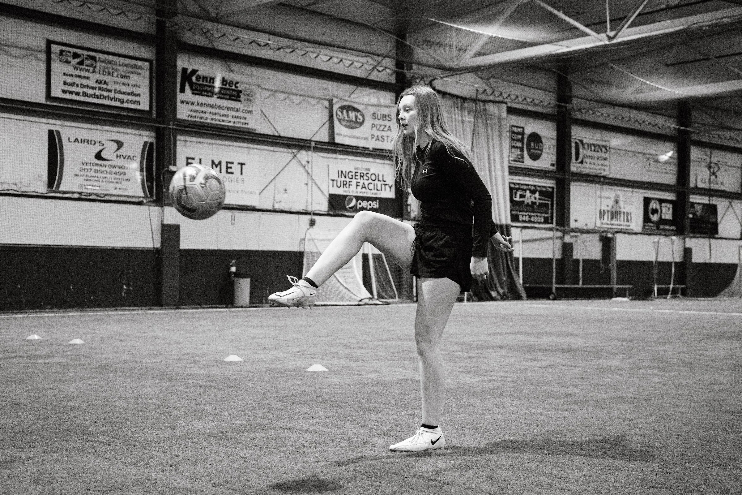 A young woman practicing soccer in an indoor sports facility, kicking a soccer ball while balancing on one leg, with cones on the field and advertisements on the wall in the background.