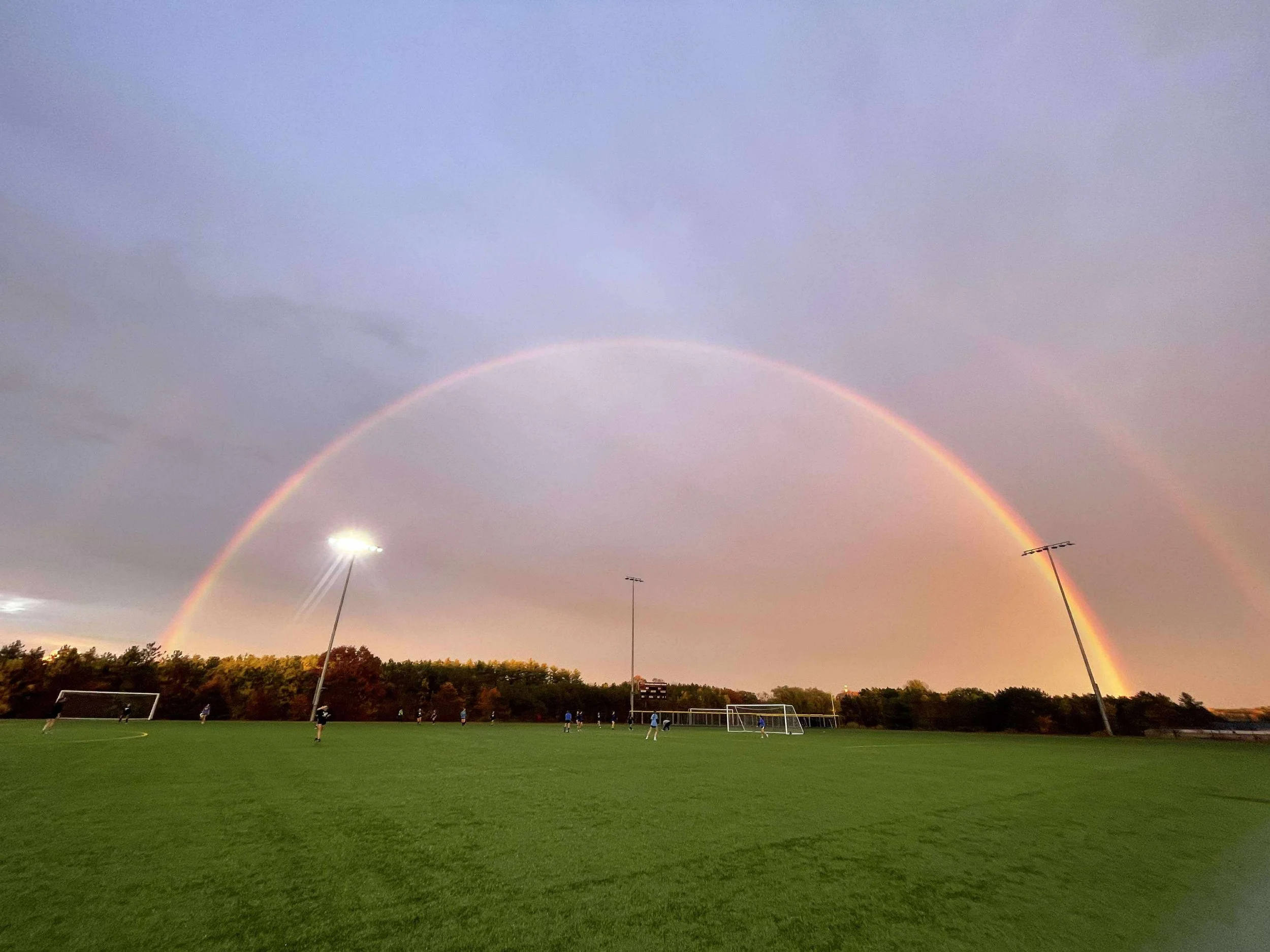 A rainbow arching across the sky over a soccer field with players and tall stadium lights.