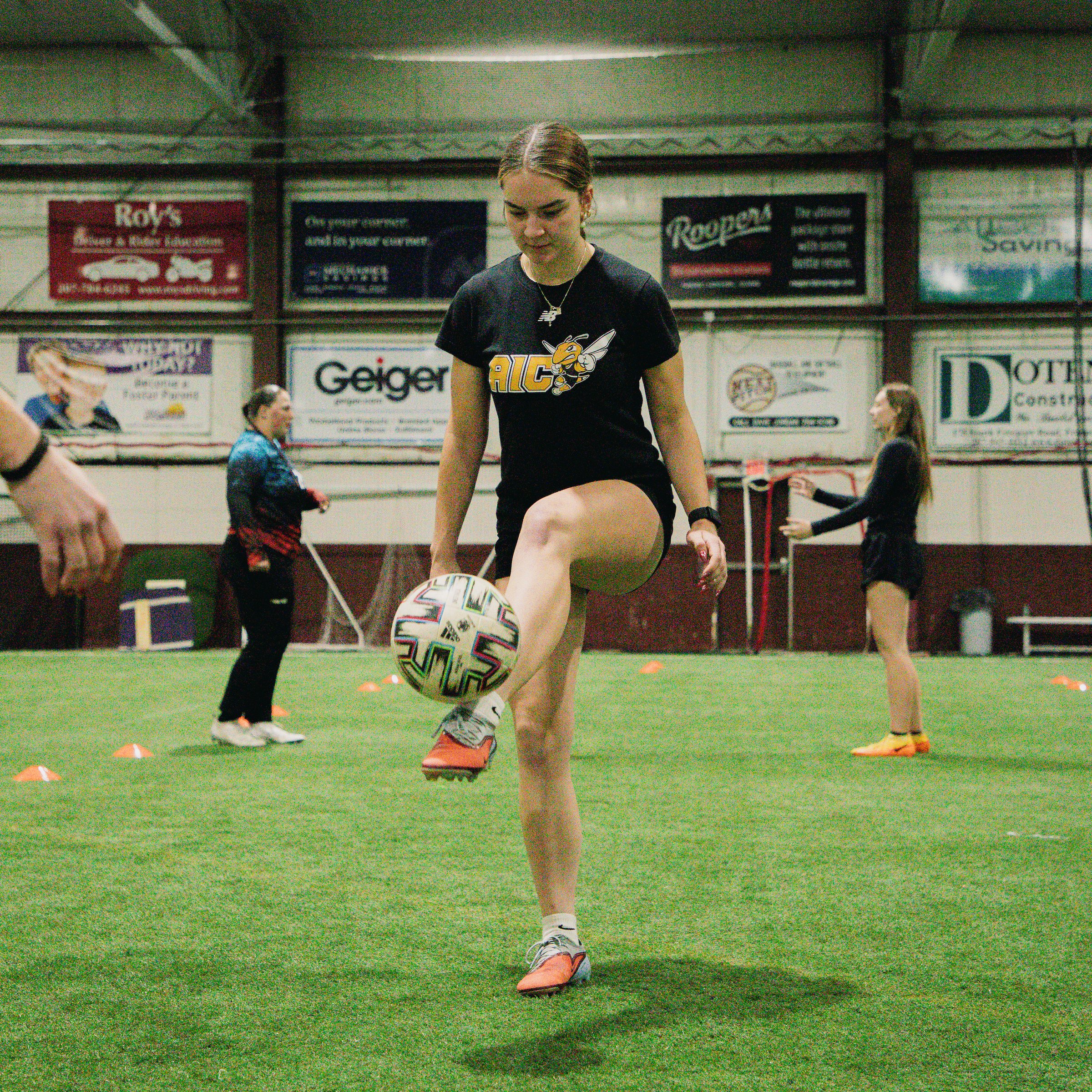 A female athlete practicing soccer on an indoor field, wearing a black t-shirt with a bee logo and black shorts, controlling a soccer ball with her foot.