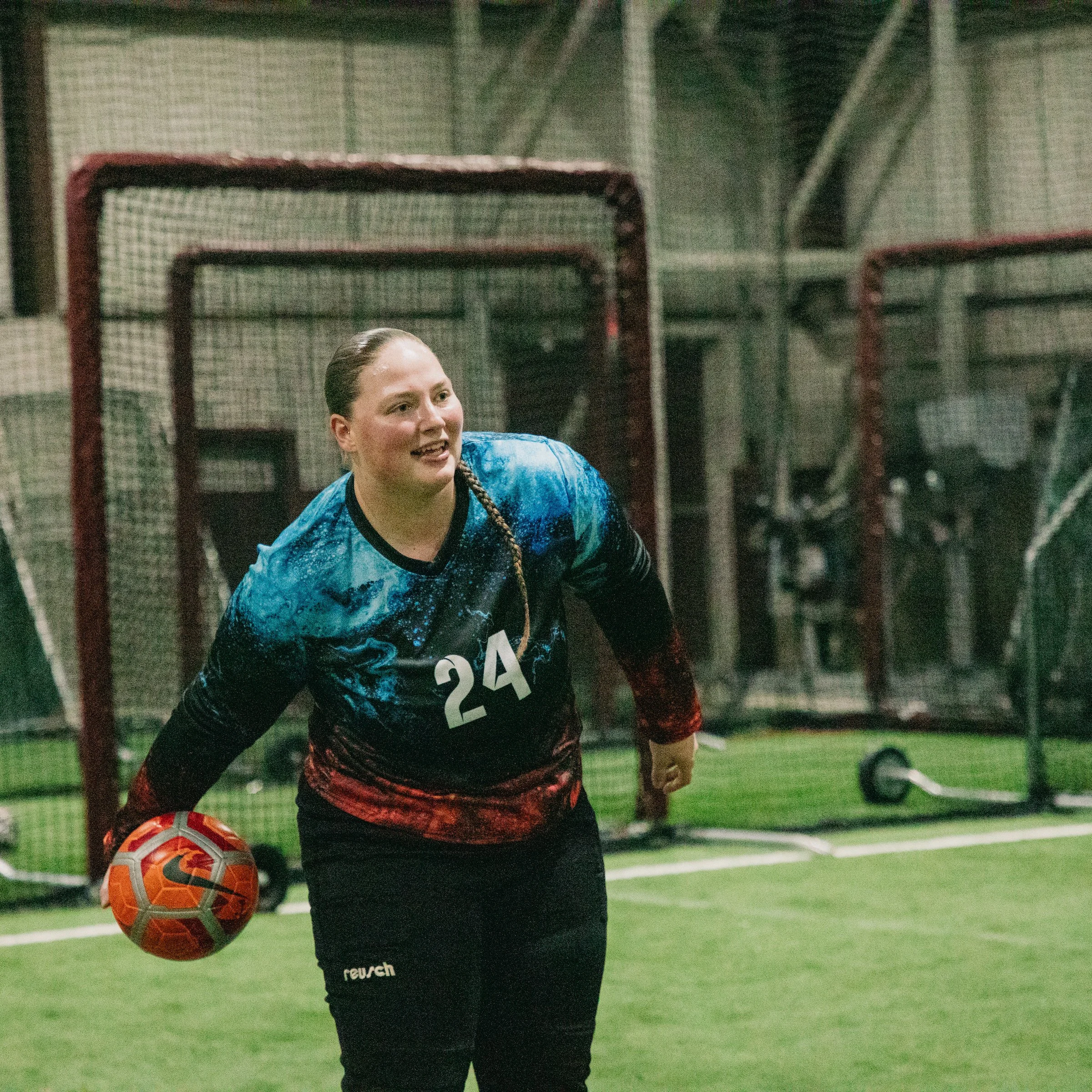 A female soccer player wearing a blue and black jersey with the number 24, holding a soccer ball inside an indoor soccer field.