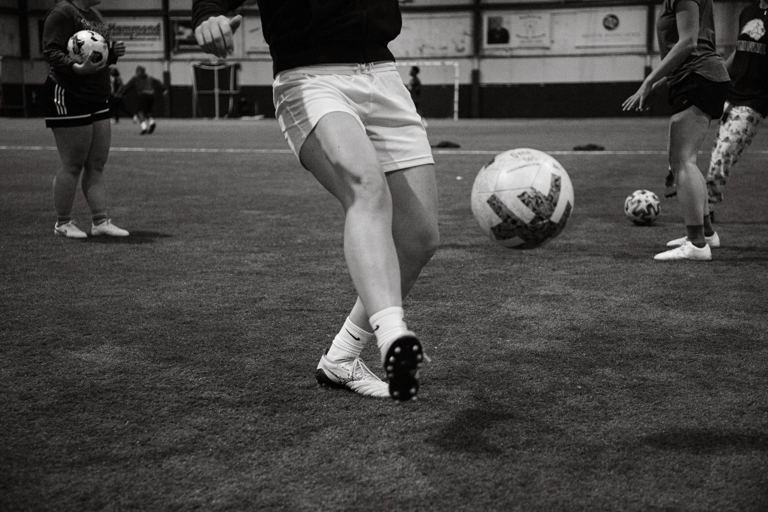 People practicing soccer indoors, with focus on a player's legs and soccer balls on the field.