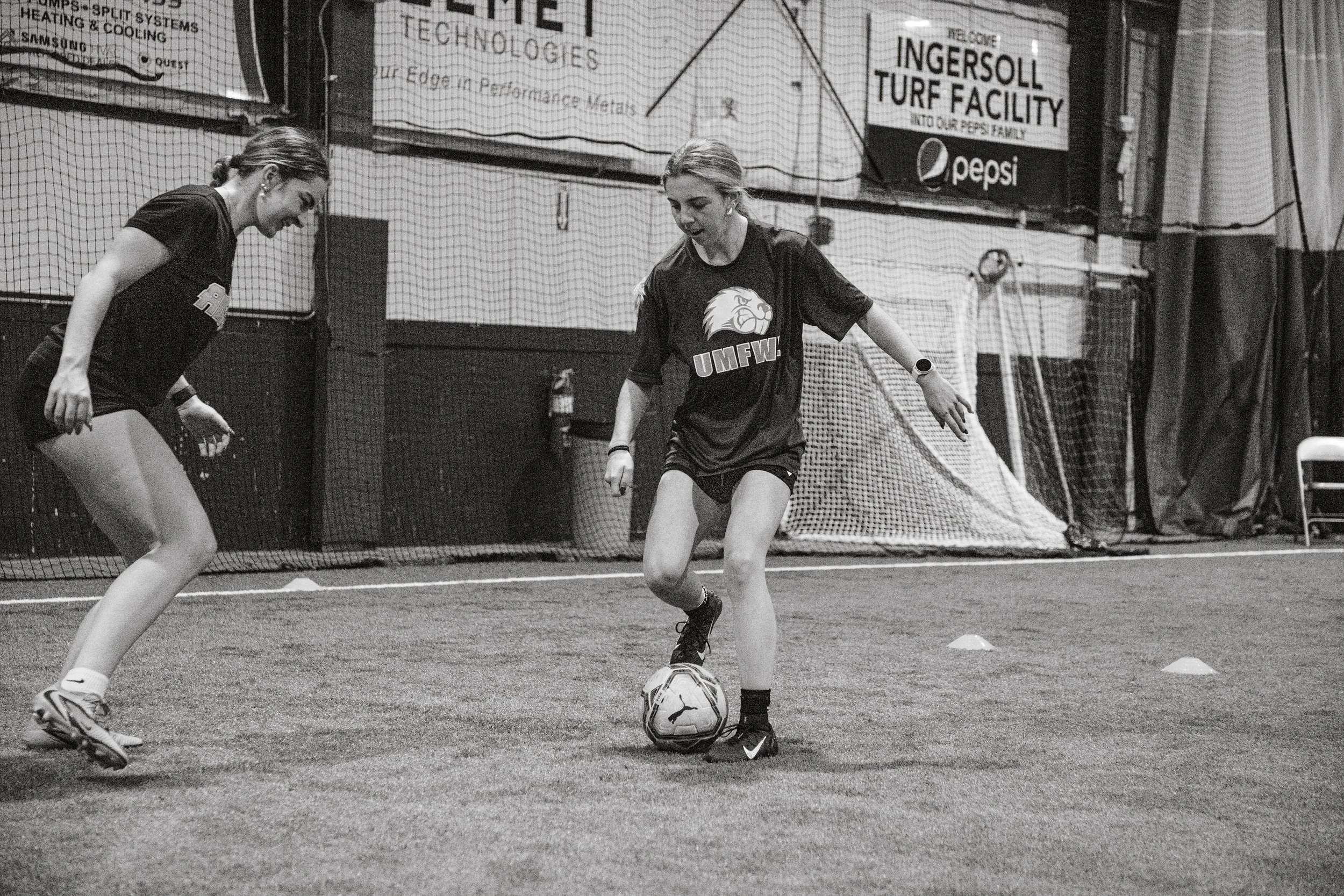 Two female soccer players practicing inside a gym. One is juggling the ball while the other observes. Both are wearing UMFW jerseys and athletic gear.
