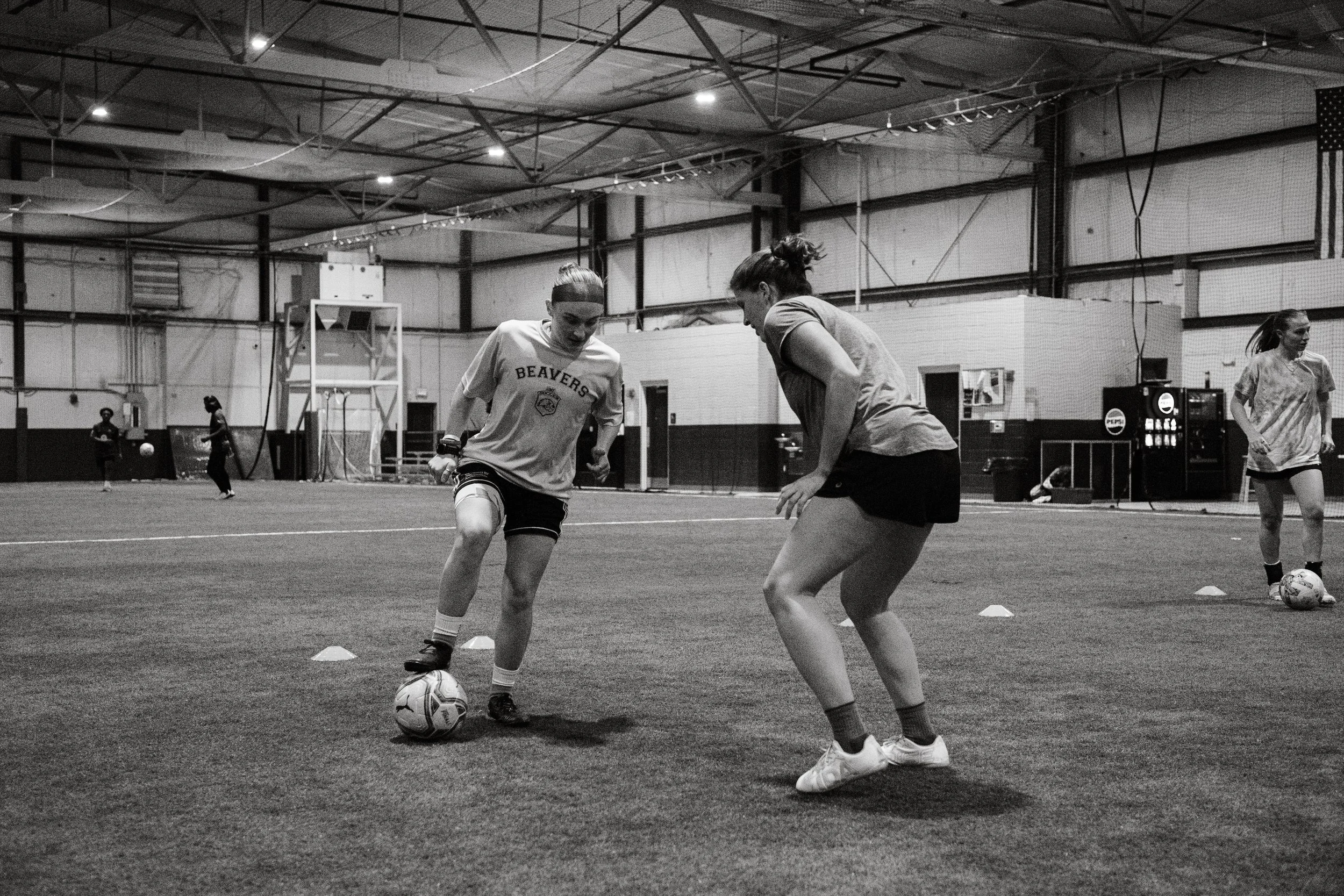Two women practicing soccer inside an indoor sports facility. One appears to be coaching or instructing the other on dribbling or ball control.