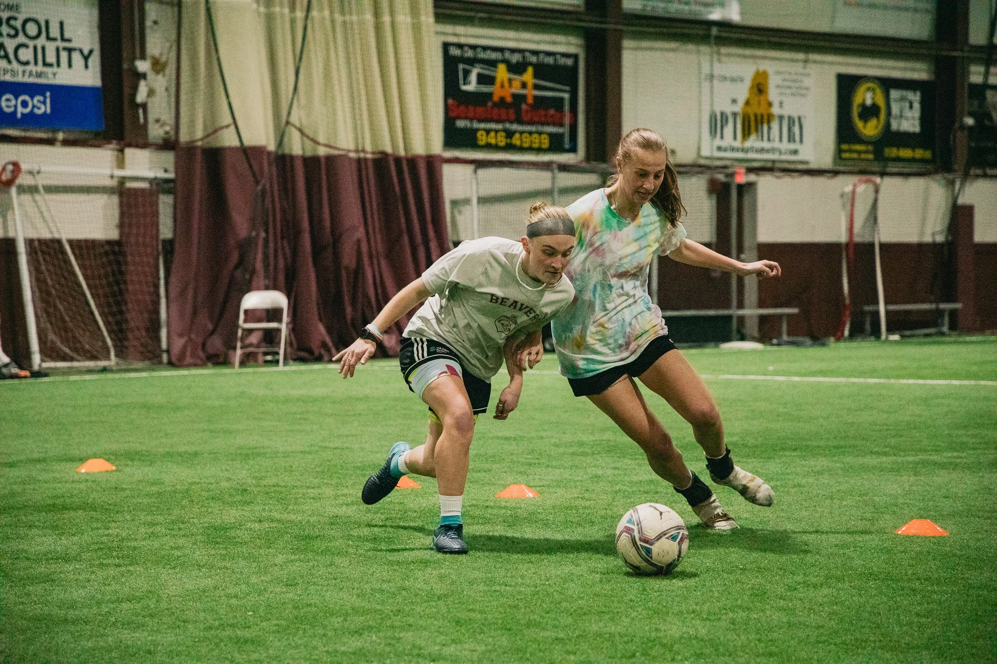 Two women playing soccer indoors, competing for the ball on a green field with orange cones nearby.