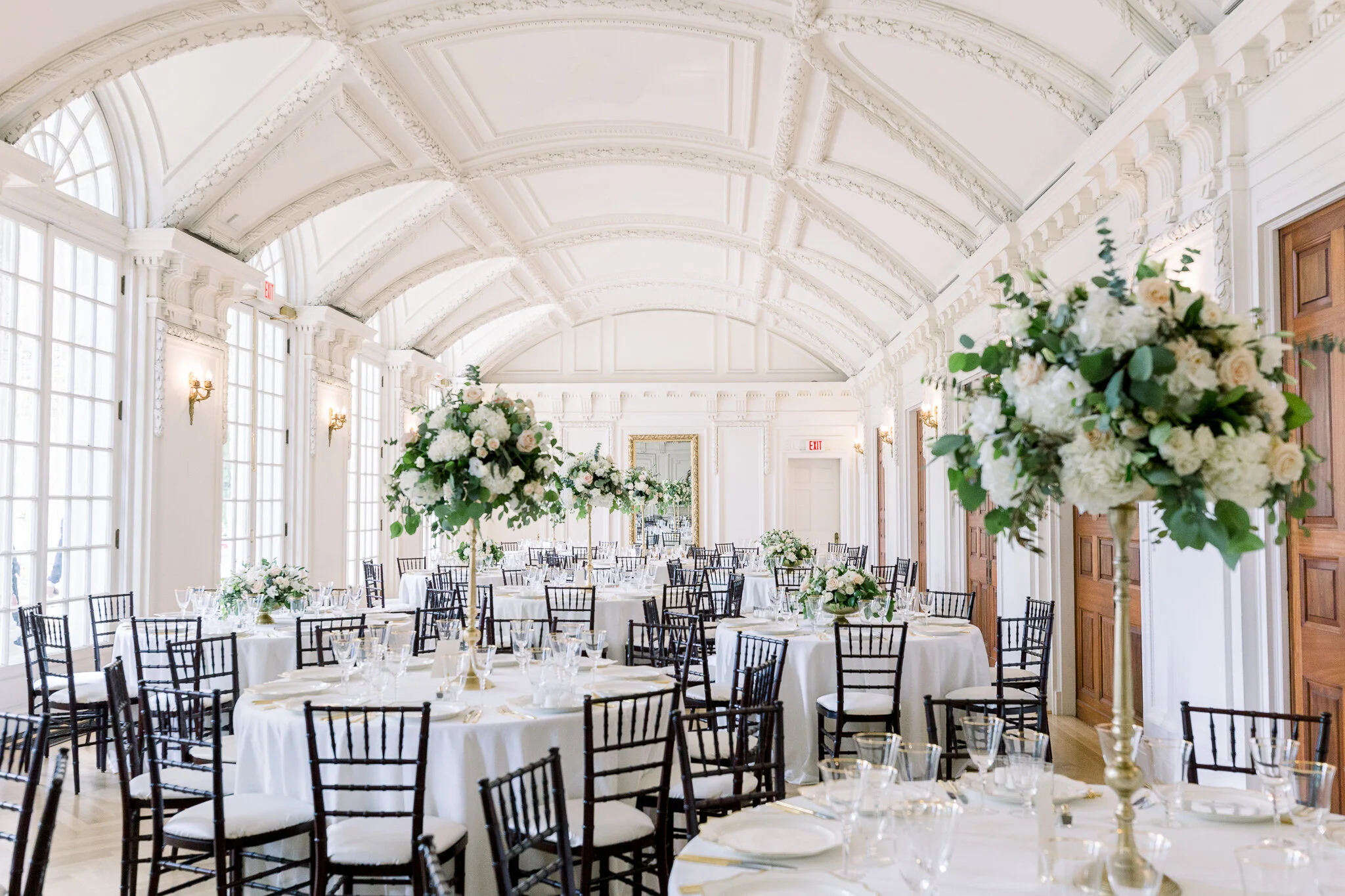 Elegant banquet hall with white walls, arched ceiling, large windows, and floral centerpieces on round tables set with white tablecloths, glassware, and silverware.