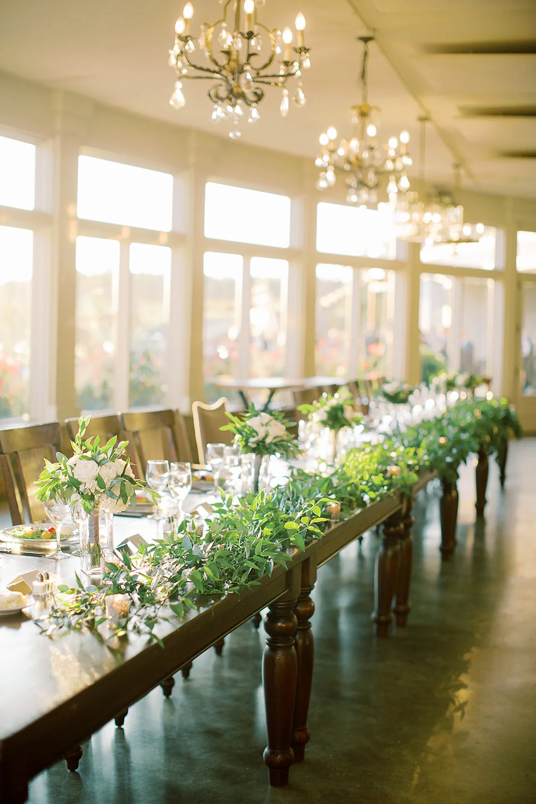 Sunlit banquet room with a long wooden table decorated with green foliage and white flowers, set for a meal with wine glasses and plates, and elegant chandeliers hanging from the ceiling.