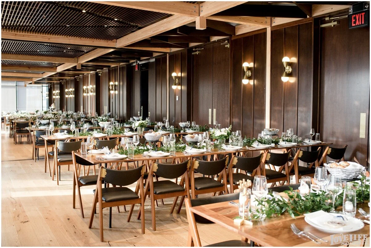 Elegant banquet hall with long wooden tables set for a formal event, decorated with greenery and white flowers, and black chairs, illuminated by wall sconces and natural light from large windows.