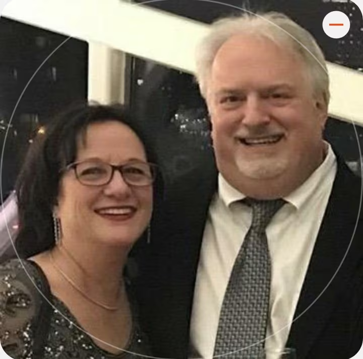 A smiling woman with dark hair, glasses, and earrings standing next to a smiling man with white hair, wearing a suit and tie, indoors at night.