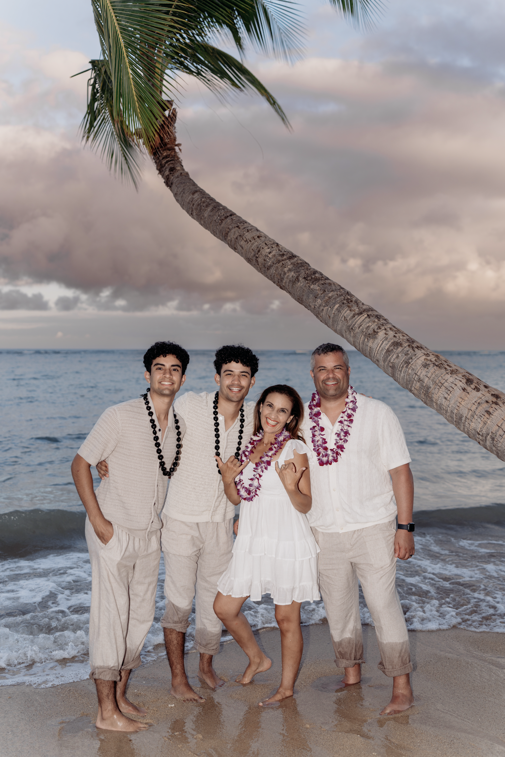 Family of four on the beach, dressed in white, with leis around their necks, standing under a leaning palm tree at sunset, smiling and enjoying the moment.