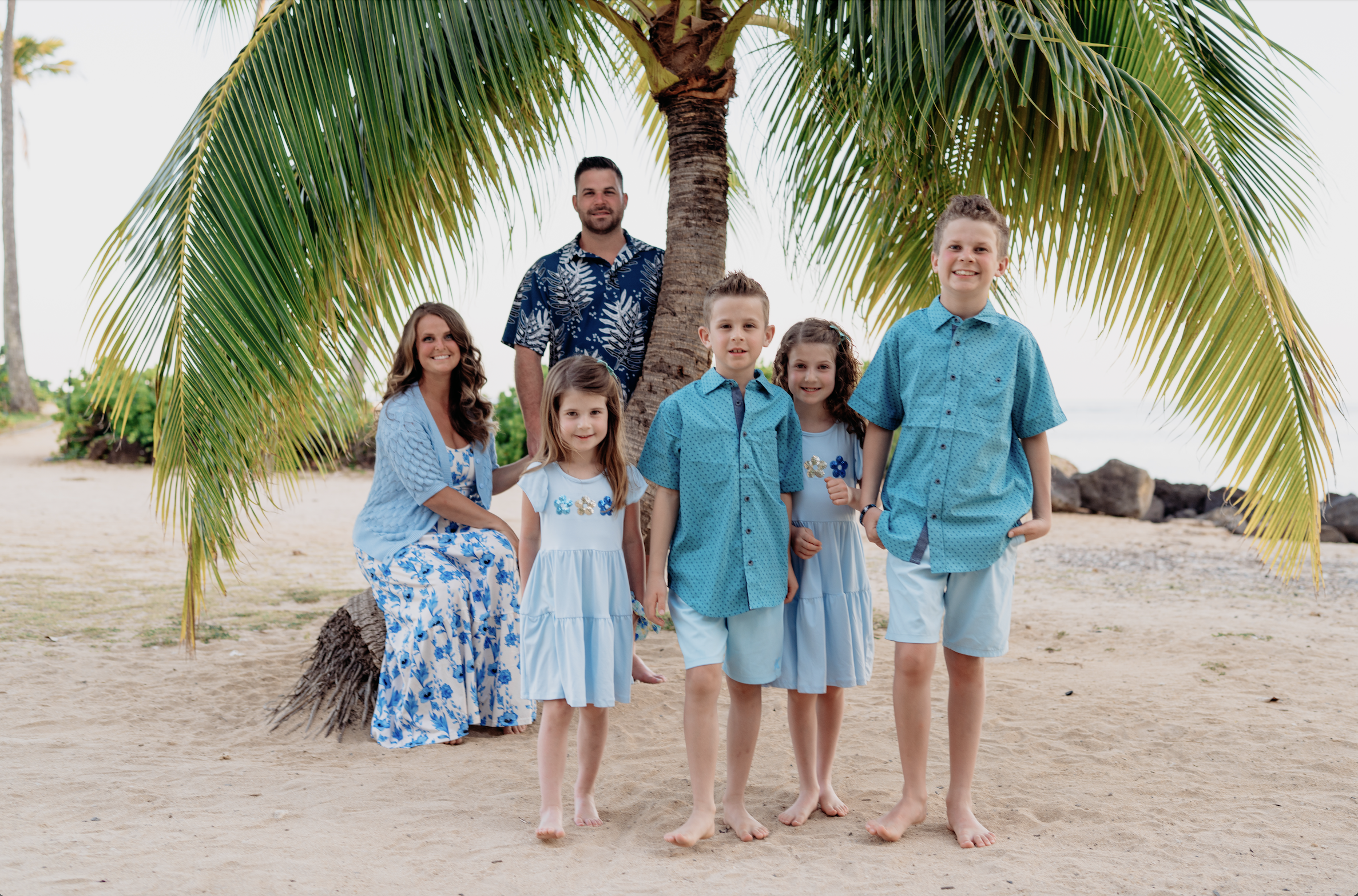 Family of seven posing on the beach under a palm tree, dressed in matching blue and white outfits.