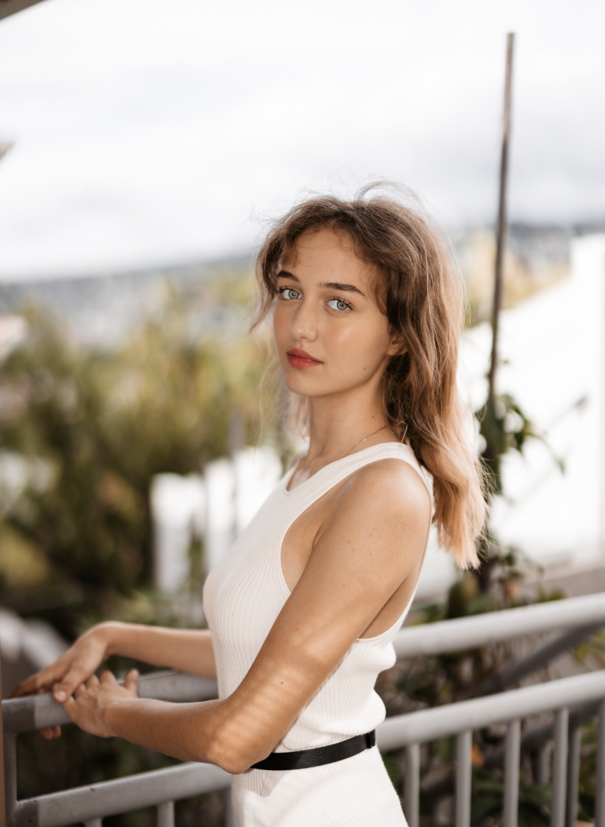 Young woman with wavy light brown hair and blue eyes standing outdoors, leaning on a railing, wearing a sleeveless white top, with blurred greenery and sky in the background.