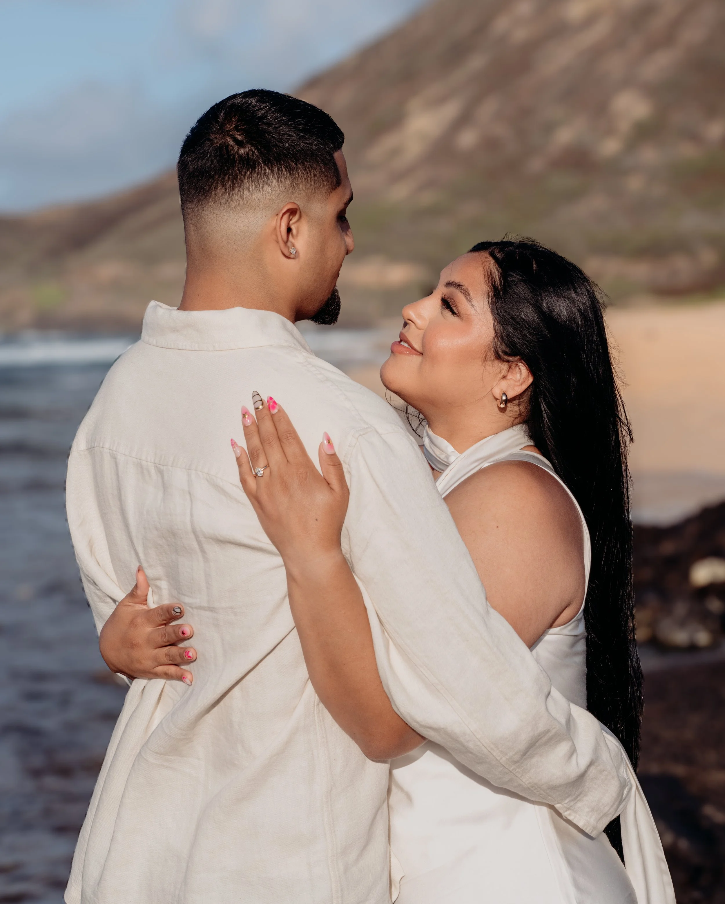 A couple hugging on the beach with mountains in the background, looking at each other affectionately.