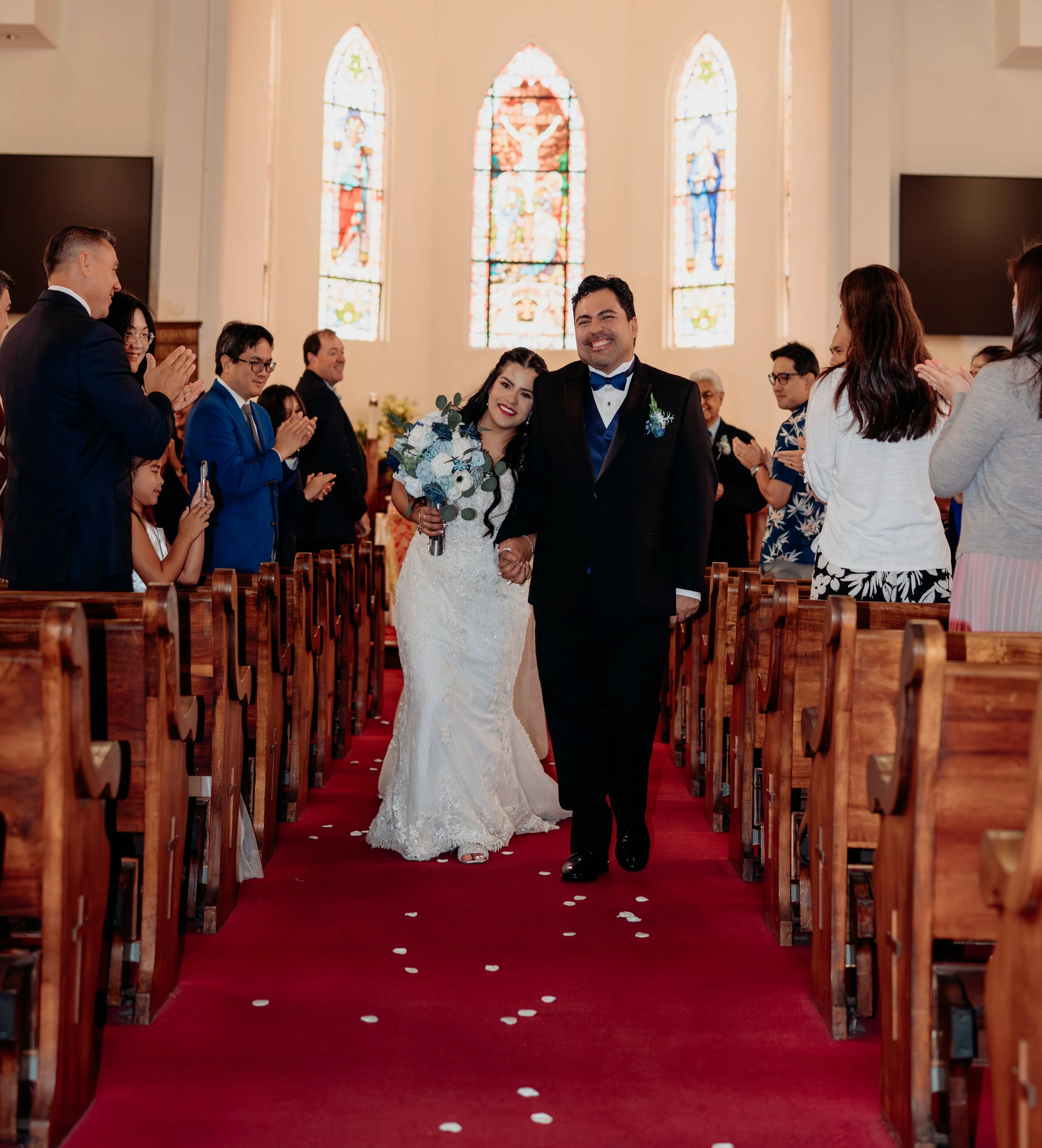 A newly married couple walking down the aisle in a church, holding hands and smiling, surrounded by friends and family.