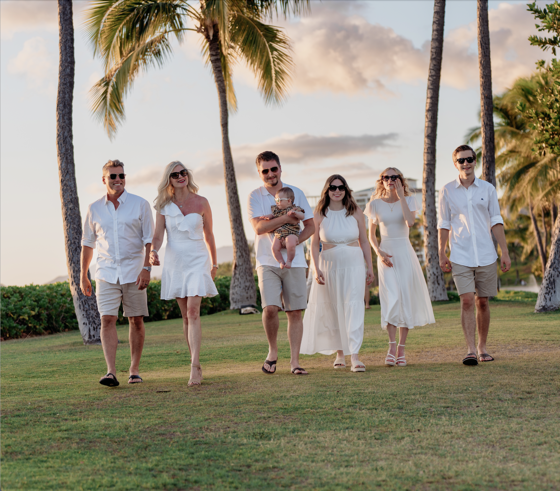A group of six adults walking together outdoors on a grassy area near palm trees, dressed in white and beige clothing during sunset.
