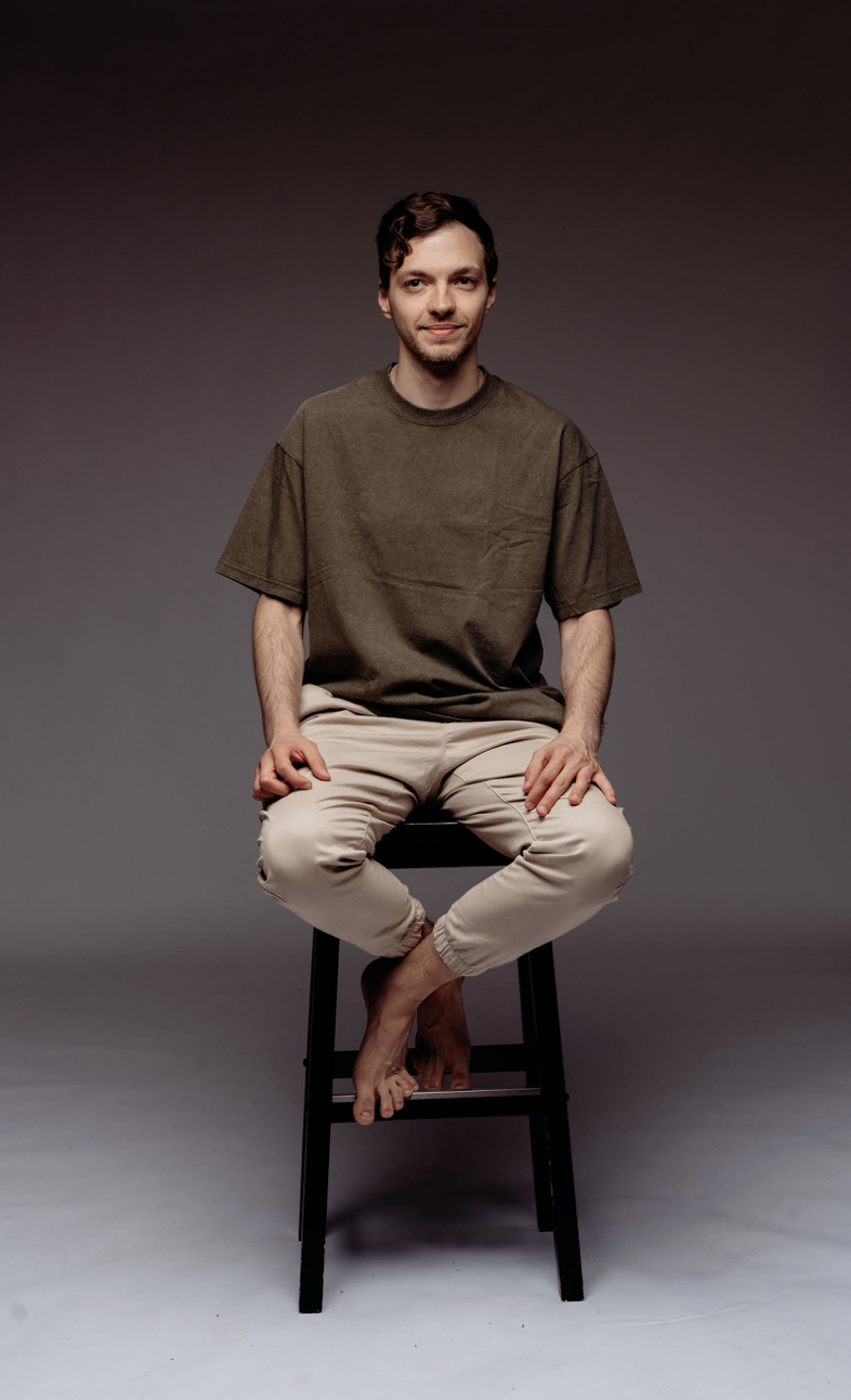 A young man sitting on a black stool with a neutral gray background. He has short wavy brown hair, a light beard, and is wearing a loose olive-green T-shirt and beige pants. He is barefoot and has a slight smile.