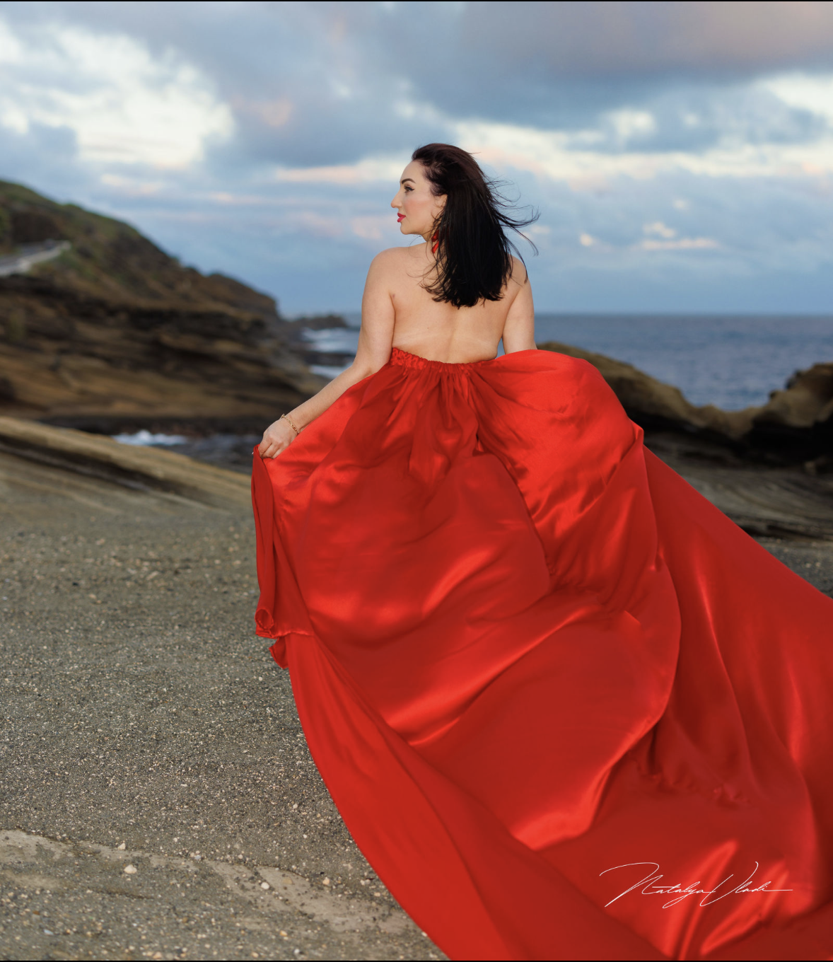 A woman in a vibrant red gown standing on a beach with rocky cliffs and the ocean in the background during cloudy weather.