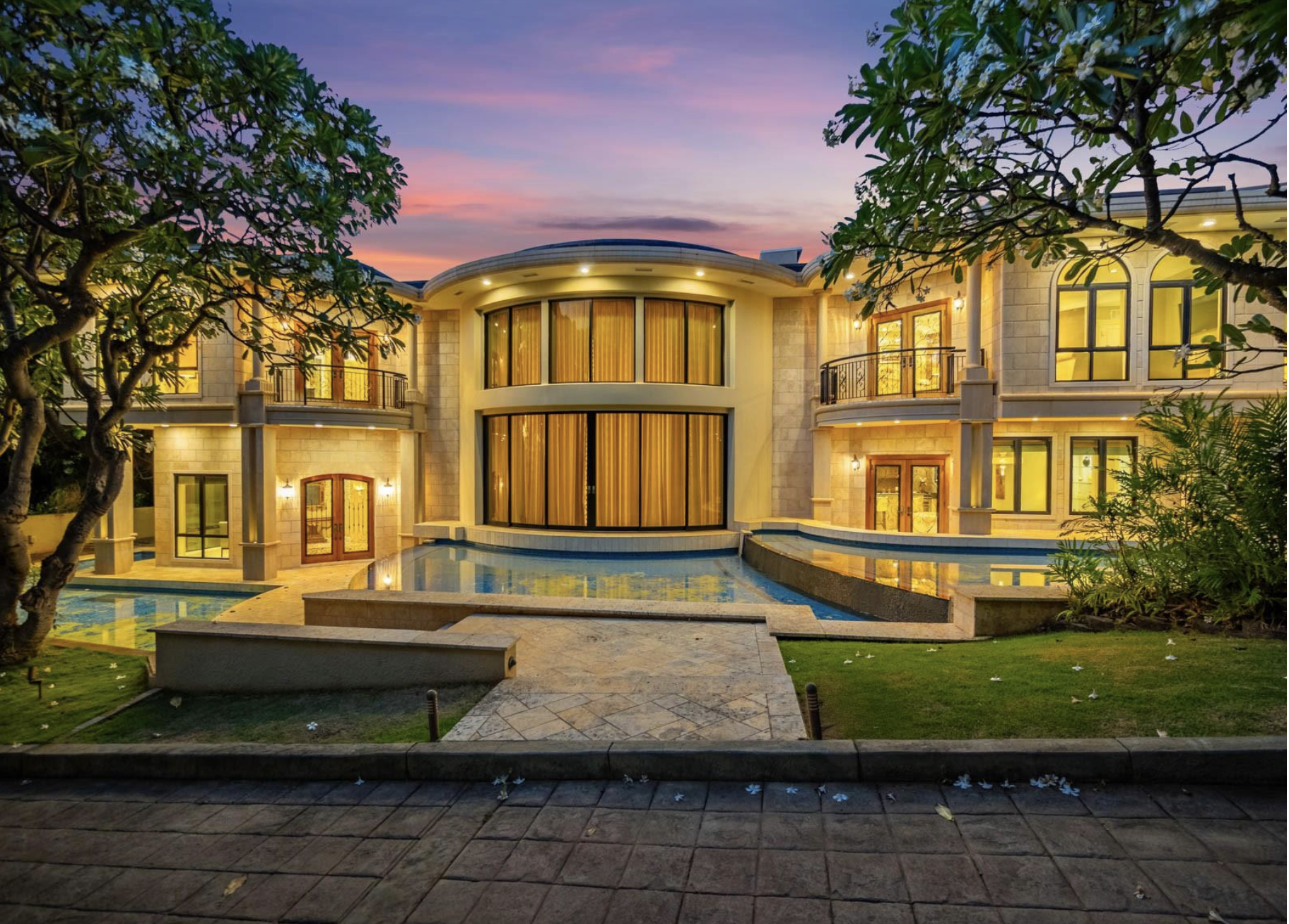 Luxury mansion with large glass windows, a water fountain, and tropical trees during sunset.