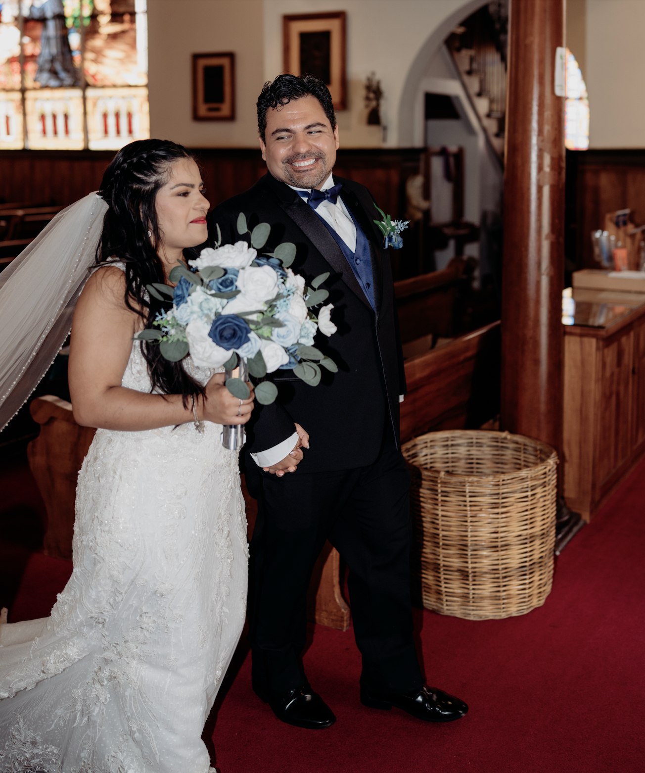 A bride and groom standing in a church with the bride holding a bouquet of white and blue flowers, both smiling.