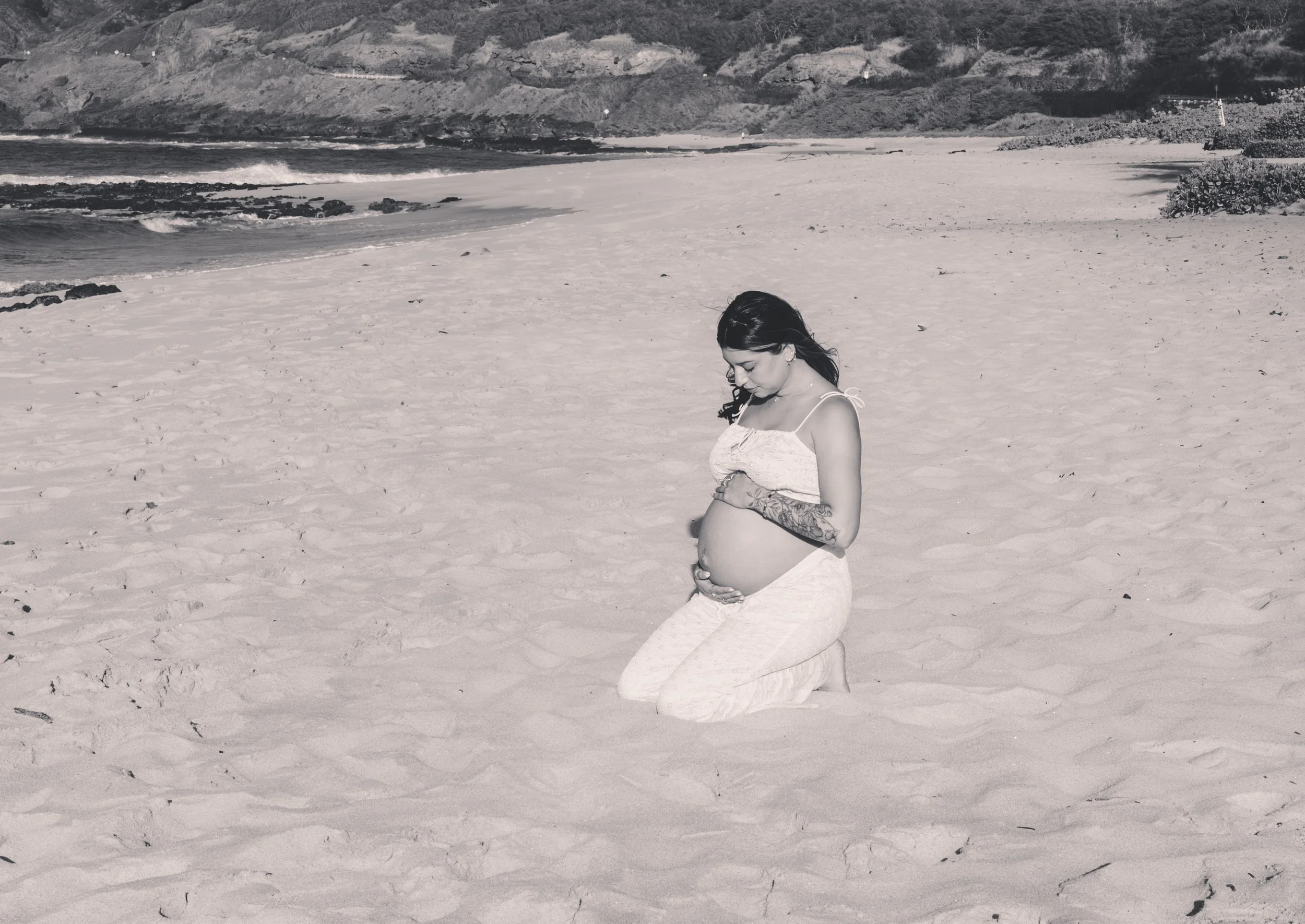 A pregnant woman with tattoos kneeling on a sandy beach, looking down and holding her belly.