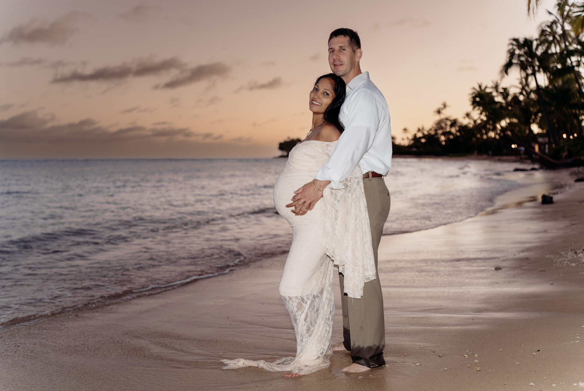 A pregnant woman in a cream-colored lace dress and a man in a white shirt and beige pants standing on a beach at sunset, with palm trees and the ocean in the background.