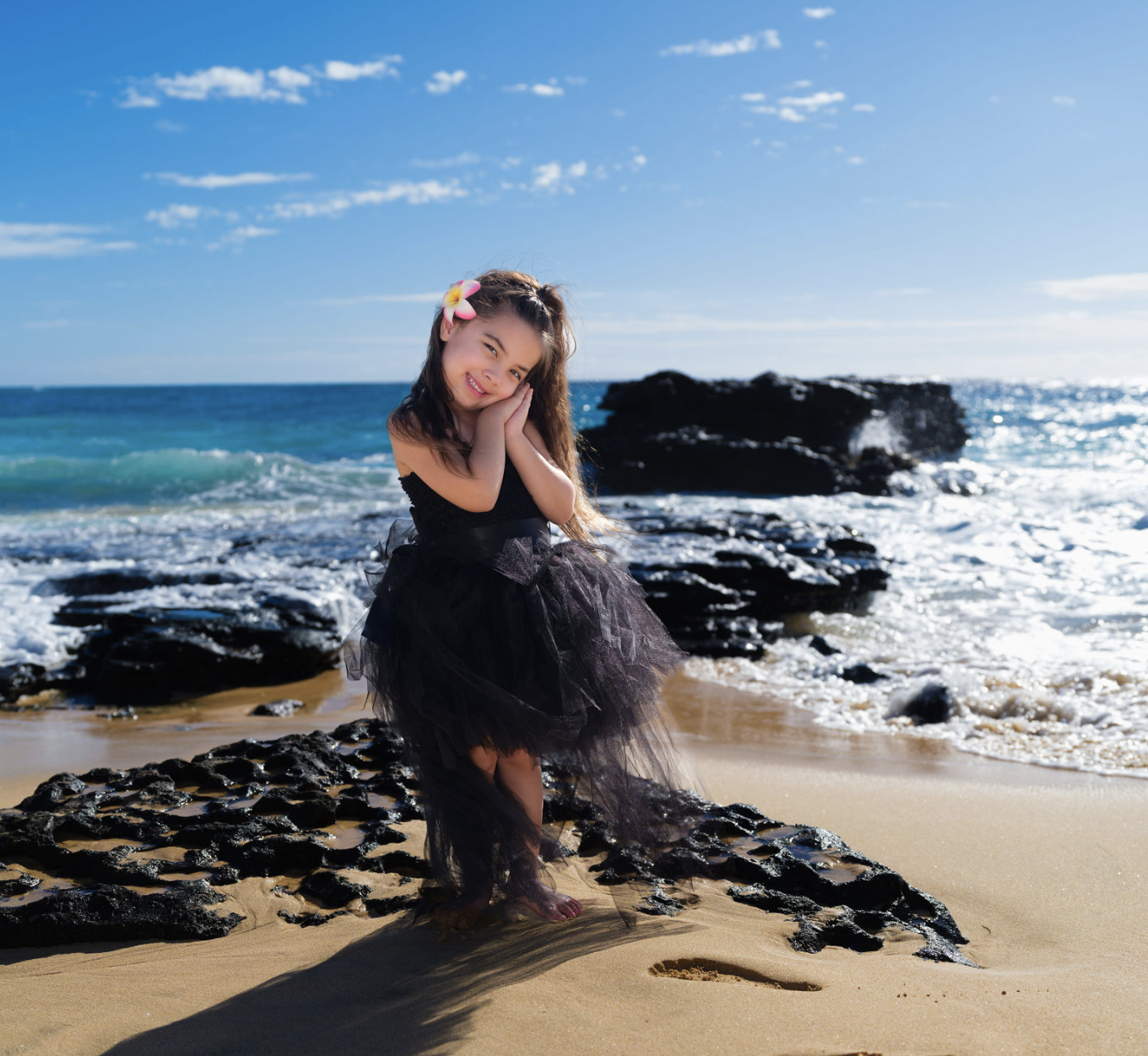 A young girl with long dark hair, wearing a black dress with a tutu skirt and a pink flower in her hair, standing on rocks at the beach, smiling with her hands folded to her cheek, with the ocean and a clear blue sky in the background.