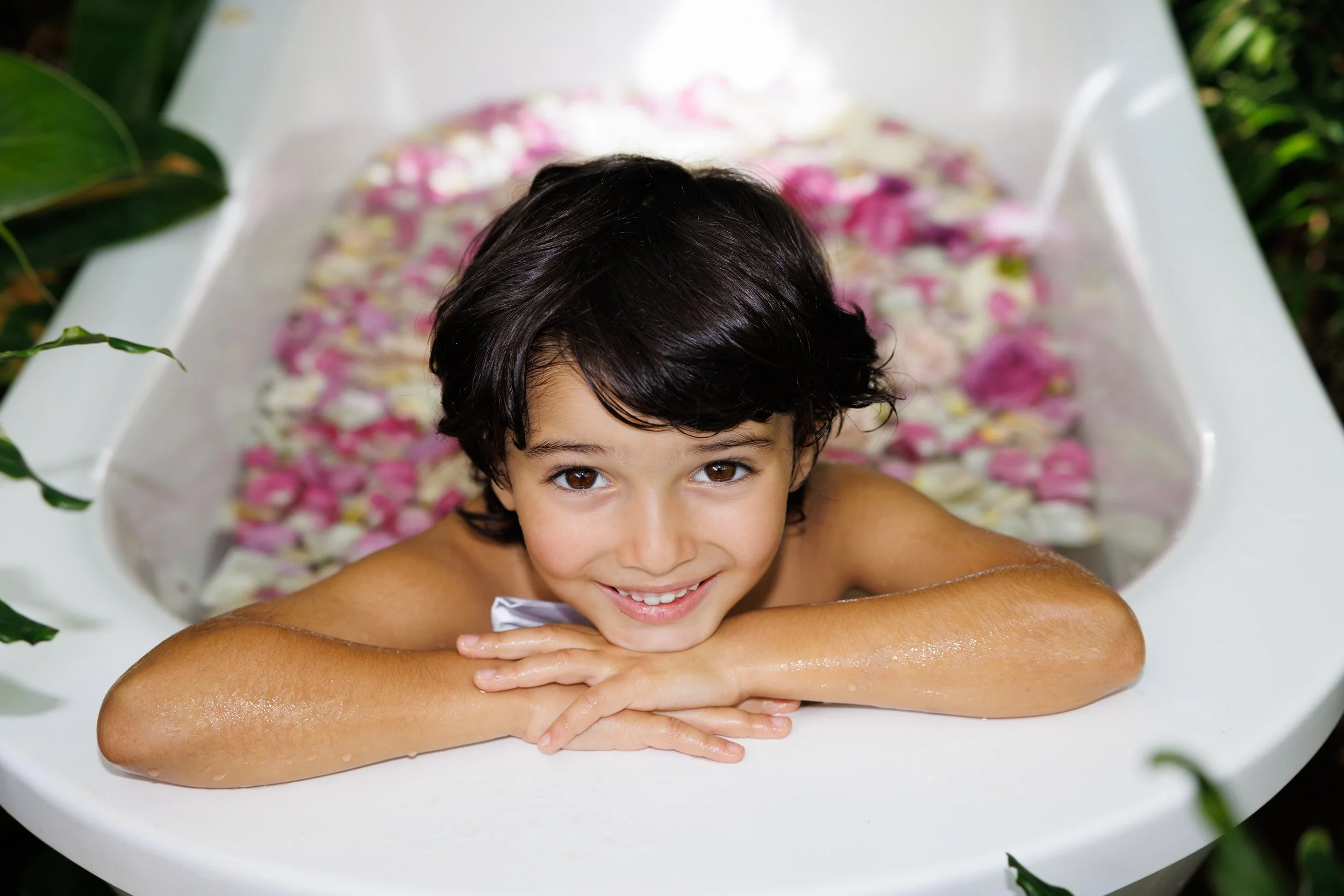 Young boy with dark hair relaxing in an outdoor bathtub filled with pink and white flower petals, surrounded by green foliage.