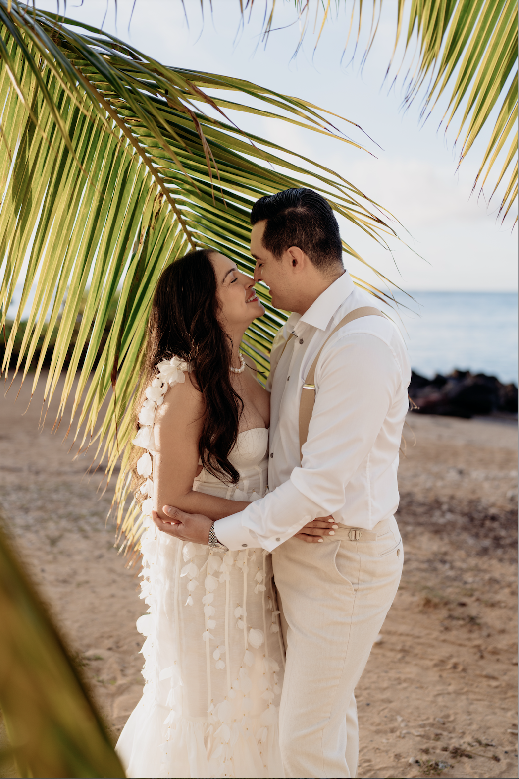 A couple embracing on a beach, with palm leaves overhead, sharing a romantic moment.