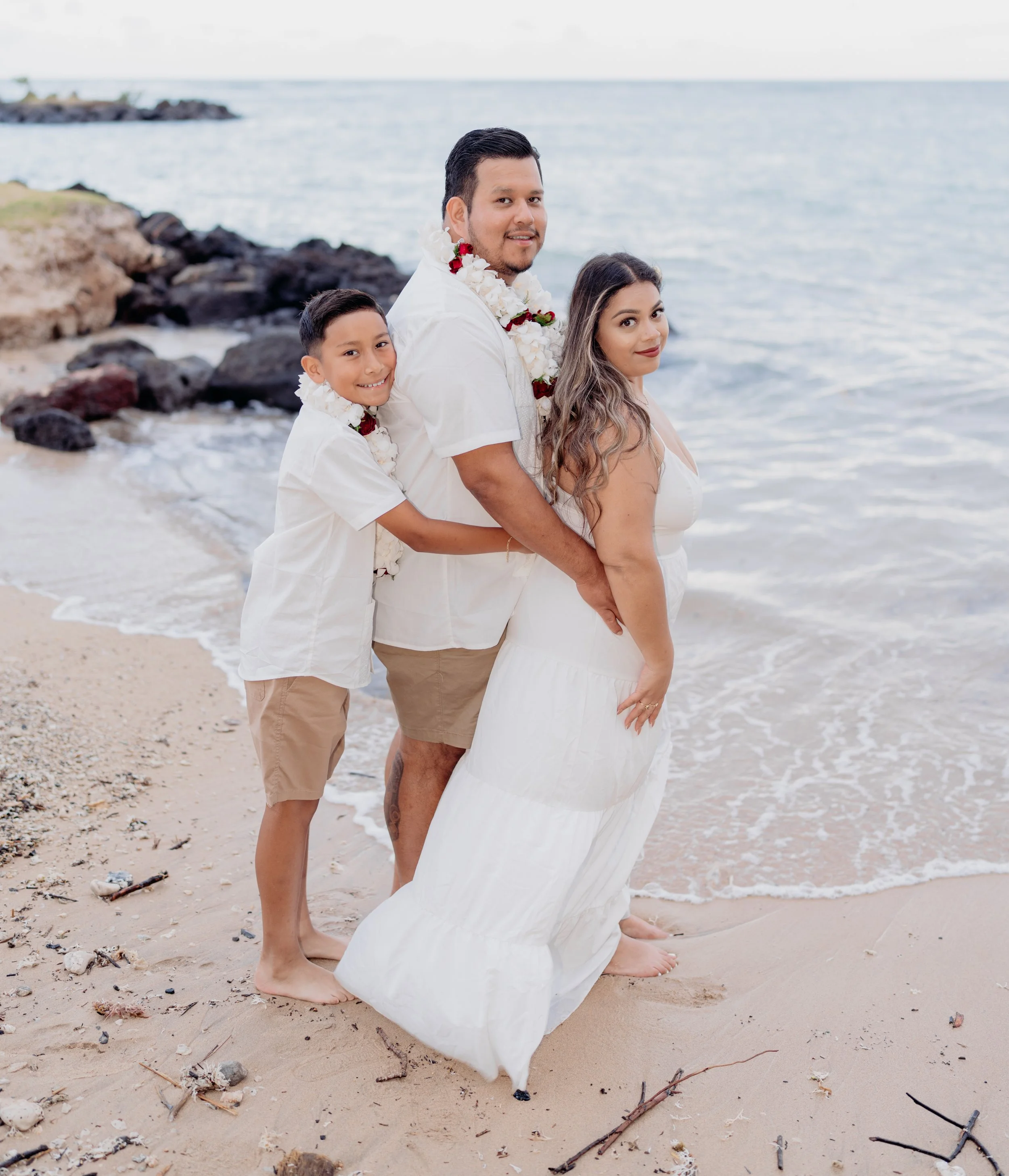 Family standing on the beach near the ocean in white clothing, with the mother in a white dress, the father and son in matching white shirts, all wearing flower leis.