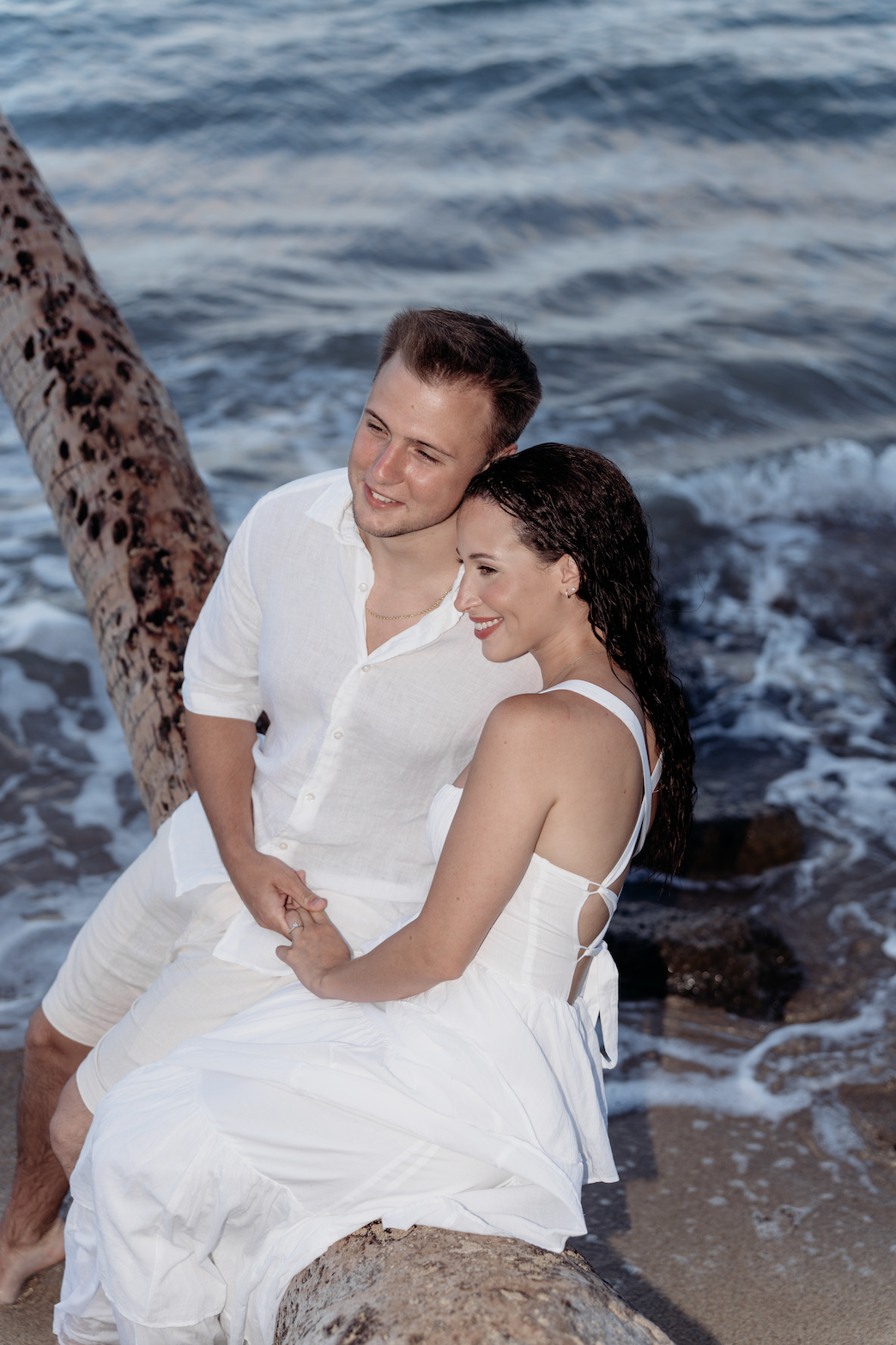 A couple dressed in white sitting on a large log on the beach near the water, smiling and enjoying the moment.