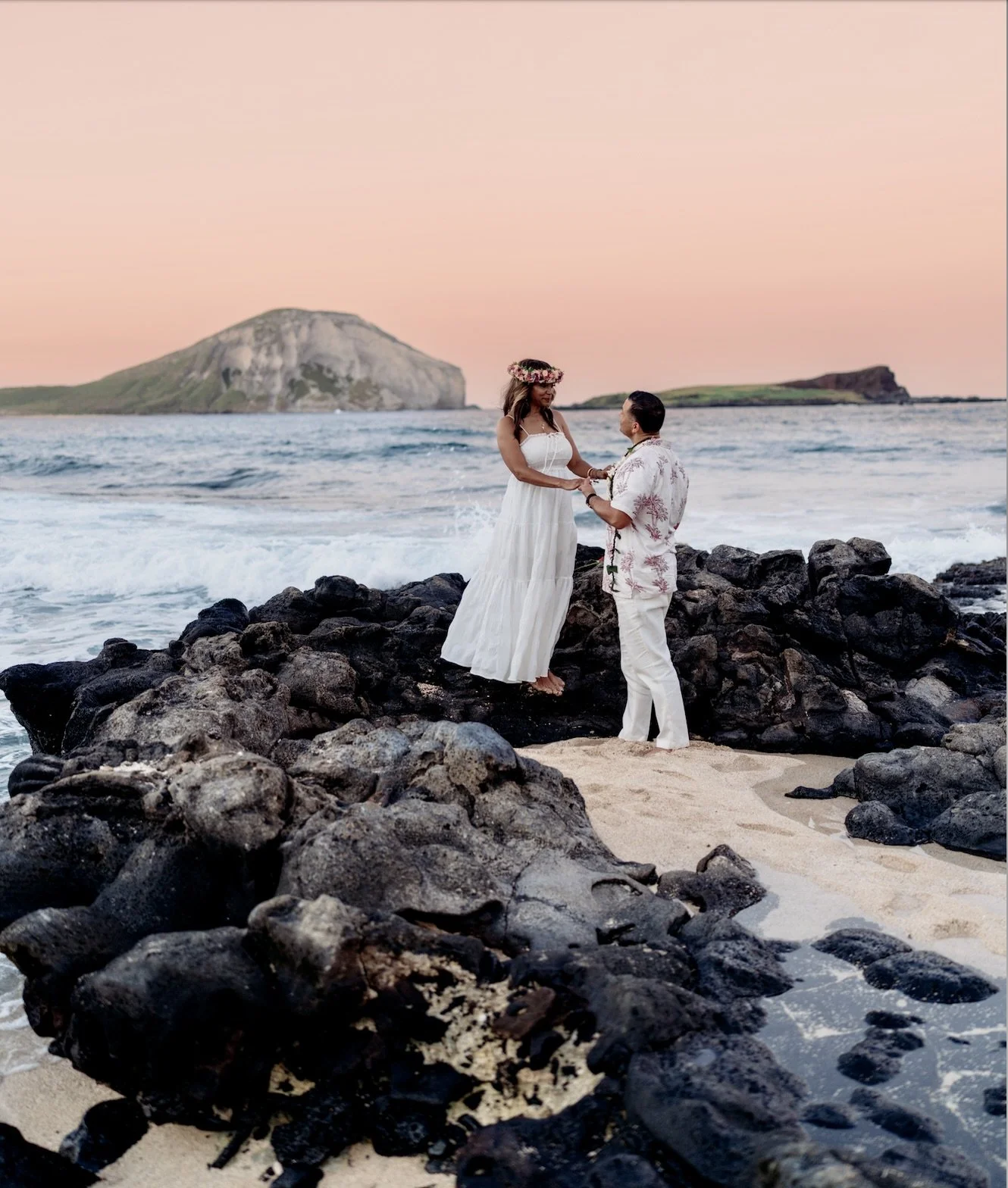 A couple getting married on a rocky beach during sunset, with the bride wearing a white dress and flower crown, and the groom in a floral shirt and white pants.