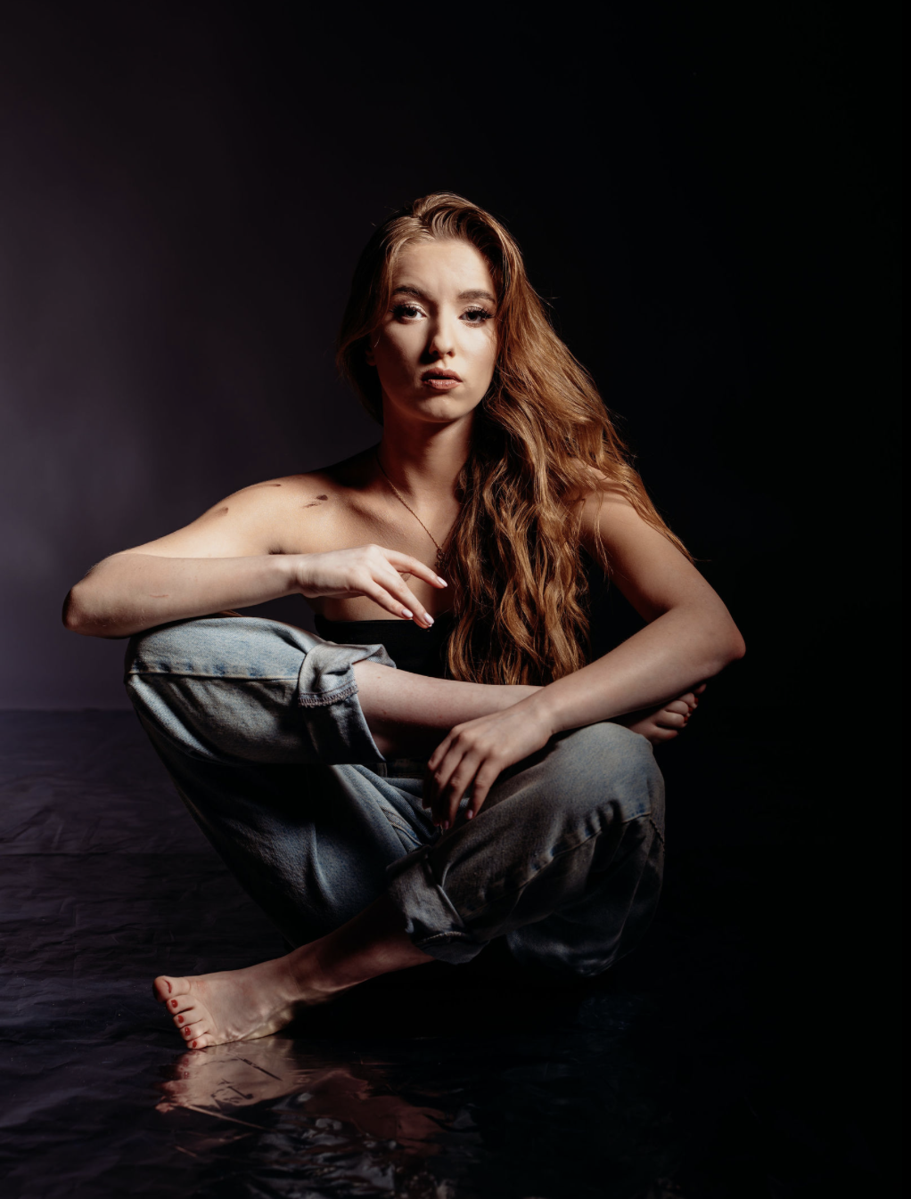 A woman with long red hair and fair skin sitting on the floor in front of a black background, wearing a black top and distressed jeans, with a serious expression on her face.