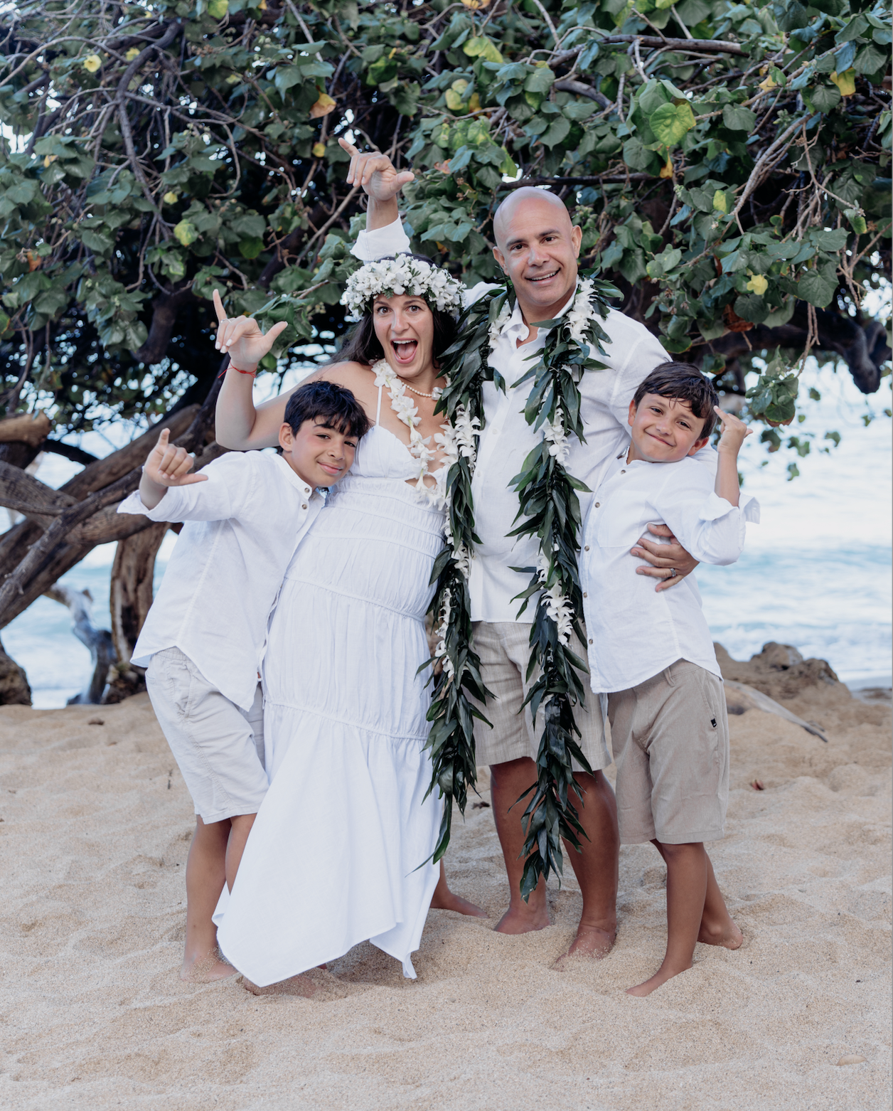 A happy family at the beach, dressed in white casual clothing, celebrating under a large leafy tree with the ocean in the background.