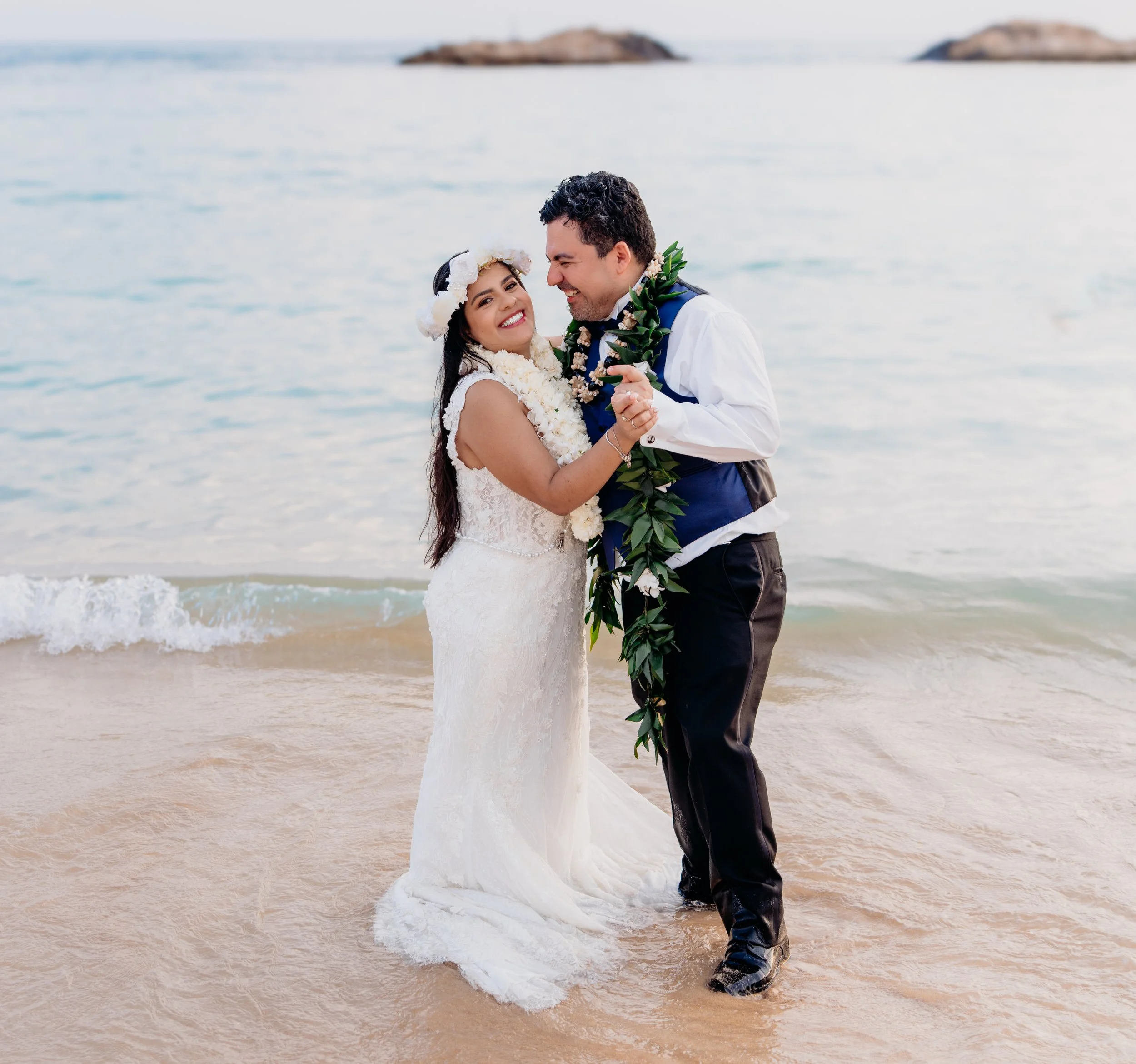 A bride and groom in wedding attire holding hands and smiling at each other on a beach with ocean backdrop.