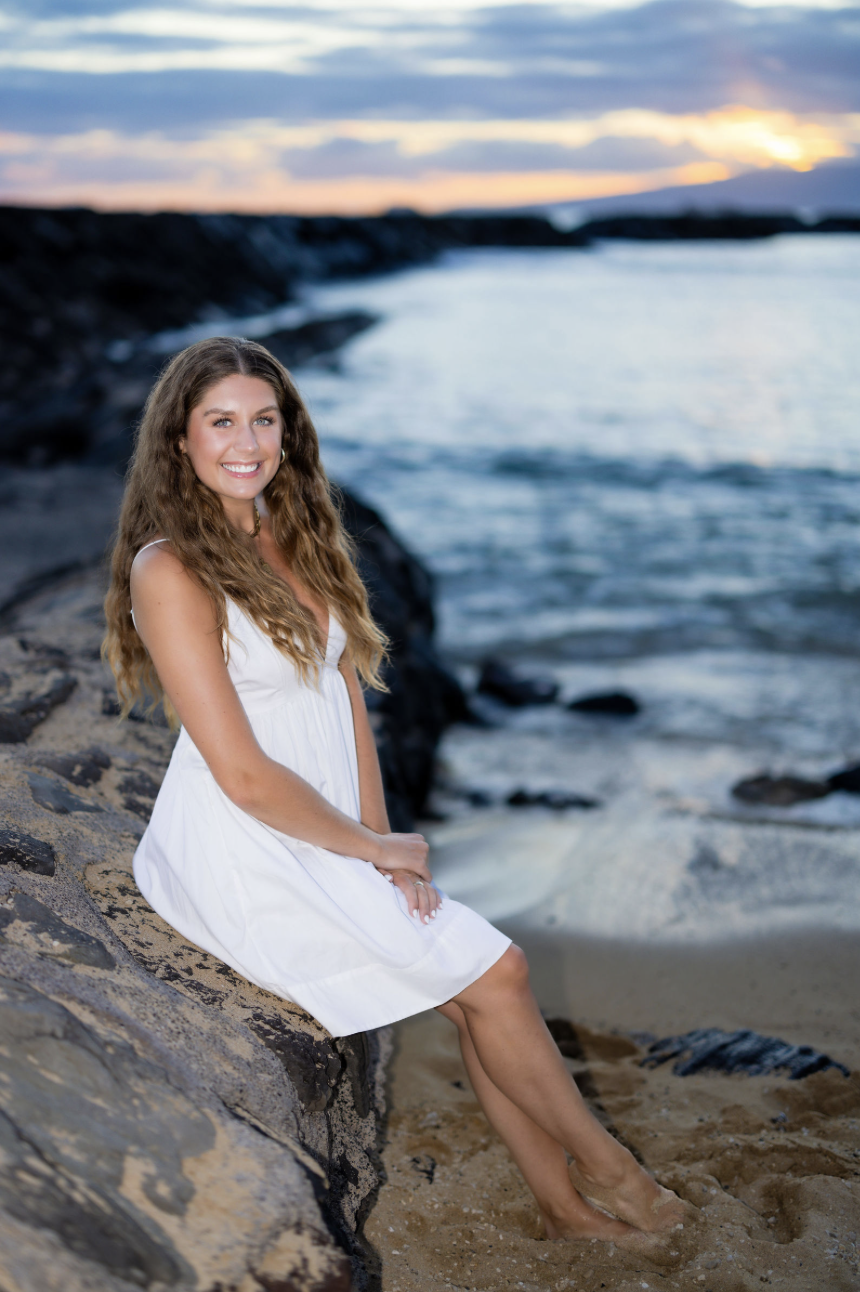A young woman in a white dress sitting on rocks by the beach at sunset, smiling at the camera
