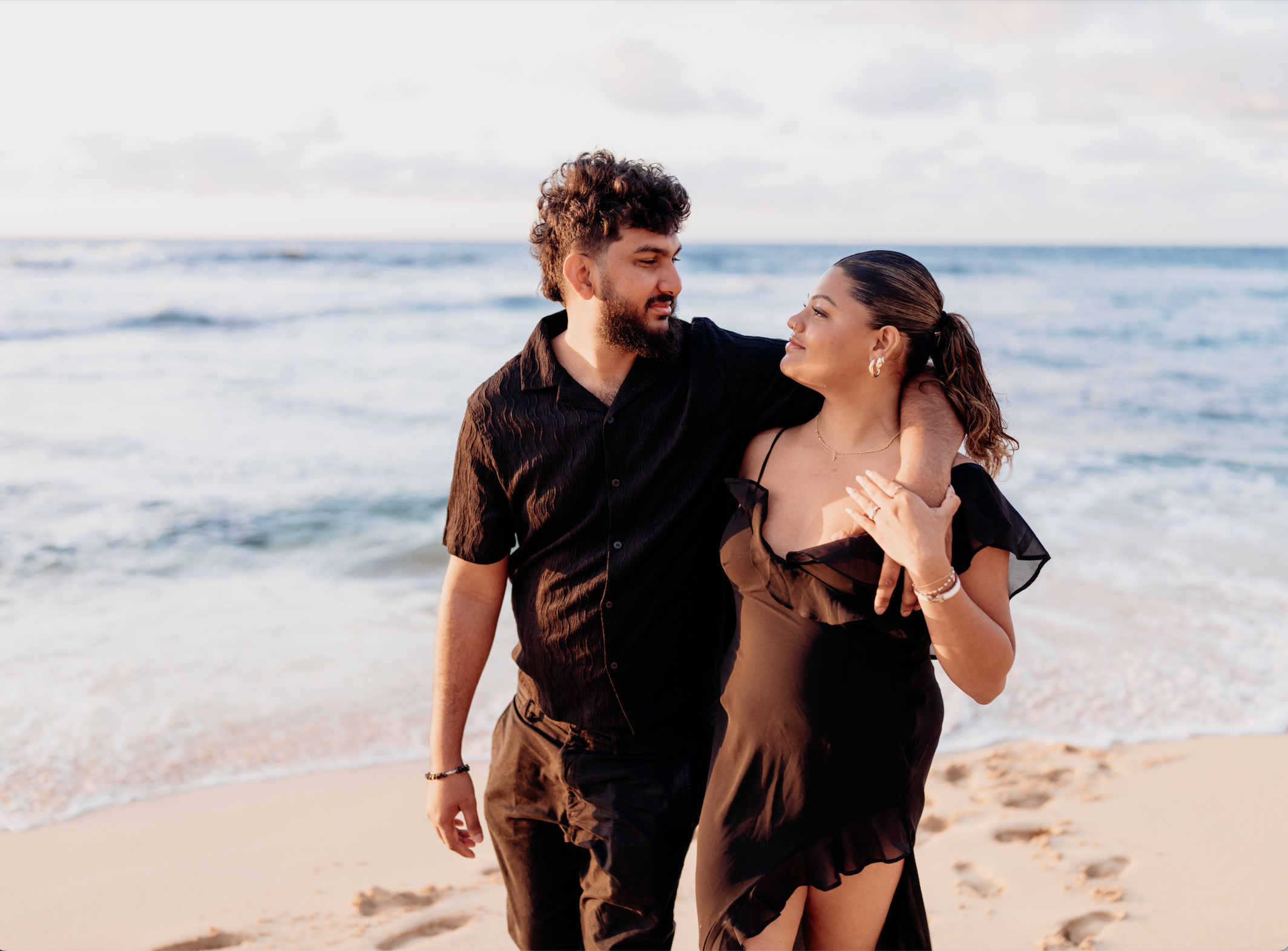 A young man and woman walk along a beach, with the ocean and cloudy sky in the background. The man has curly hair and a beard, and the woman has brown hair tied back, wearing earrings. They are smiling and look at each other affectionately, with the 