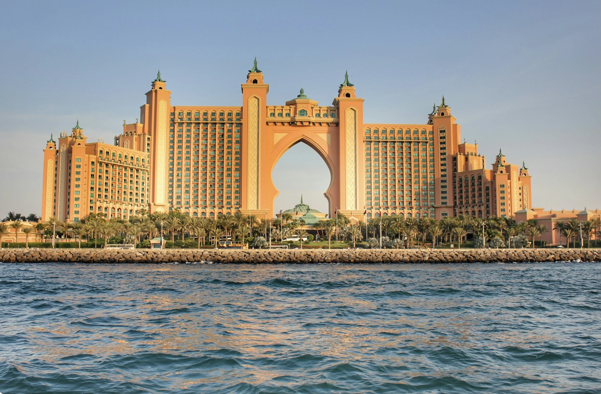 The Atlantis Paradise Island Resort in Nassau, Bahamas, viewed from the water with palm trees and a stone barrier in the foreground.