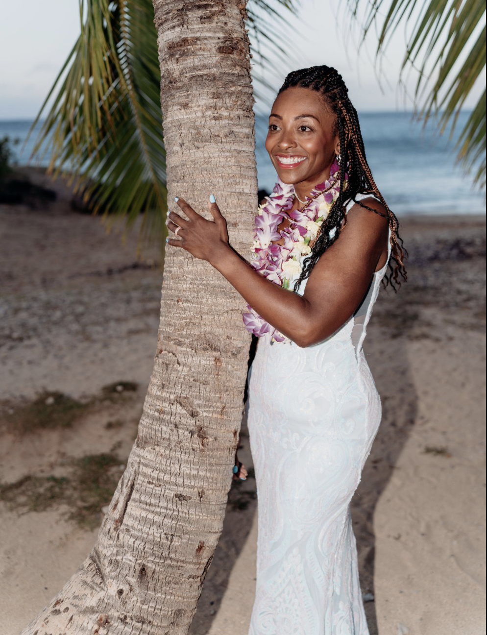 A woman with braided hair wearing a white dress and a flower lei, smiling while hugging a palm tree on a beach.