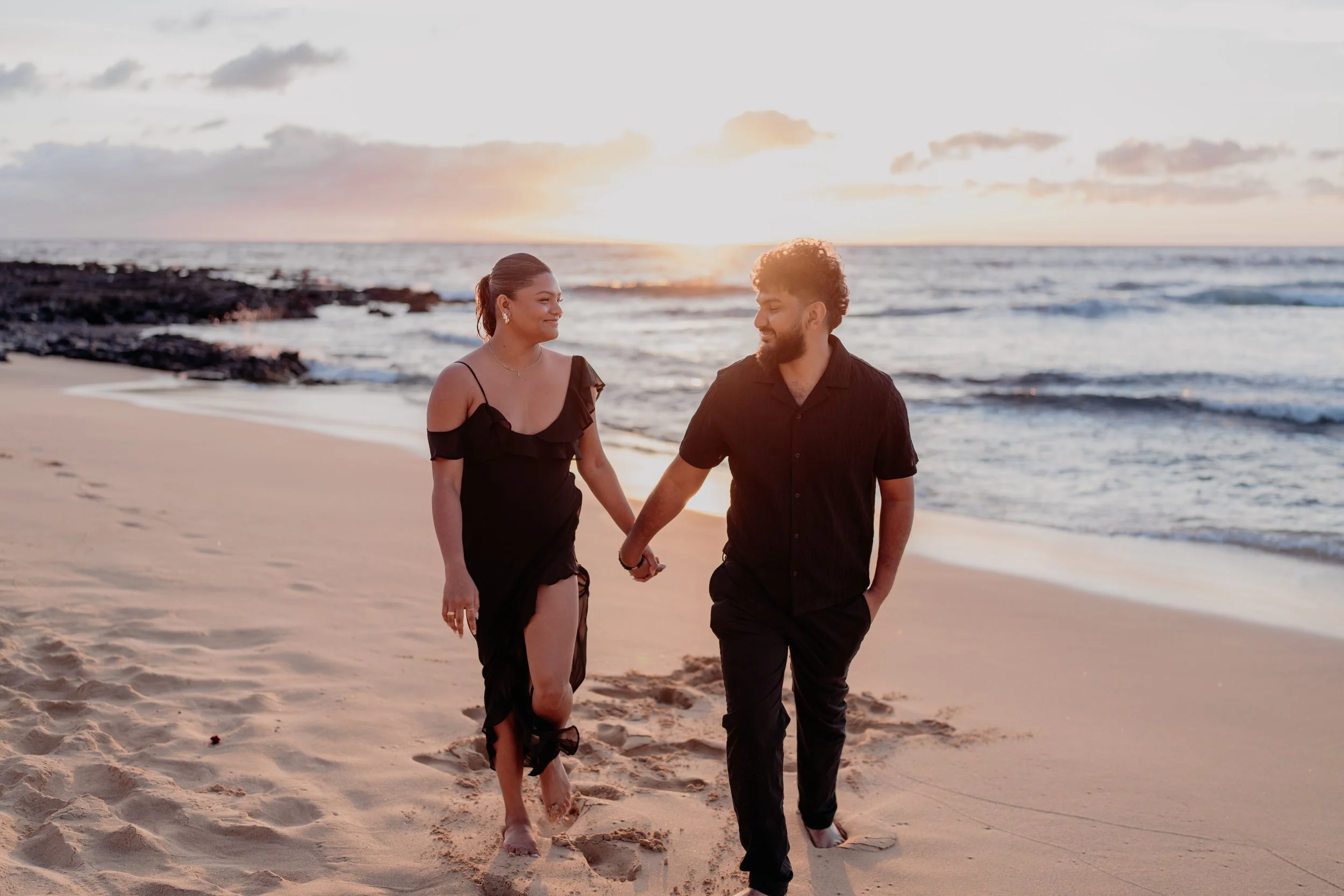 A couple walking hand in hand on a sandy beach at sunset, the woman in a black dress and the man in a black shirt and pants, with ocean waves in the background.