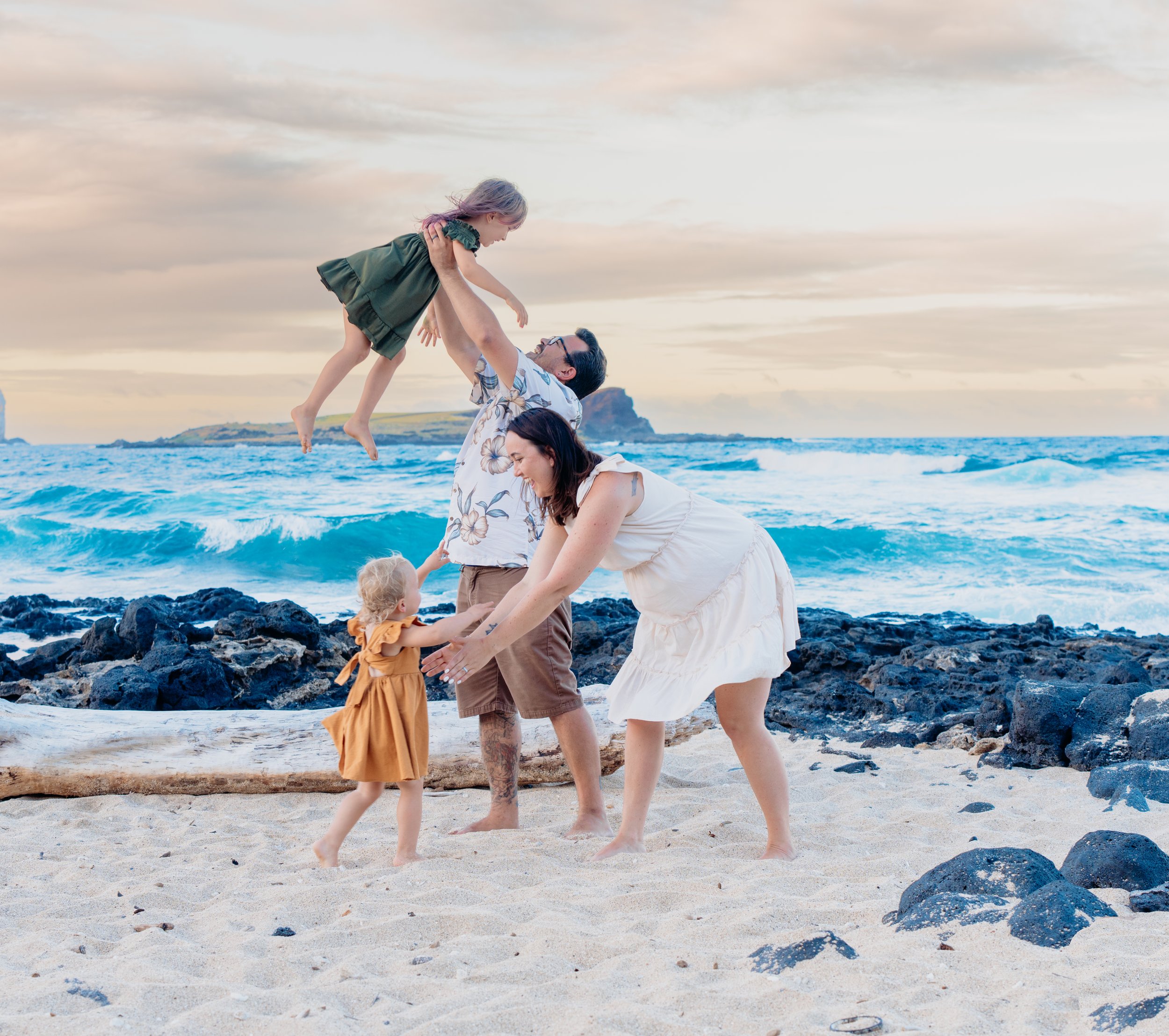 A family of five playing on a beach with rocks and ocean waves, a person lifting a young girl in the air while others smile and reach out.