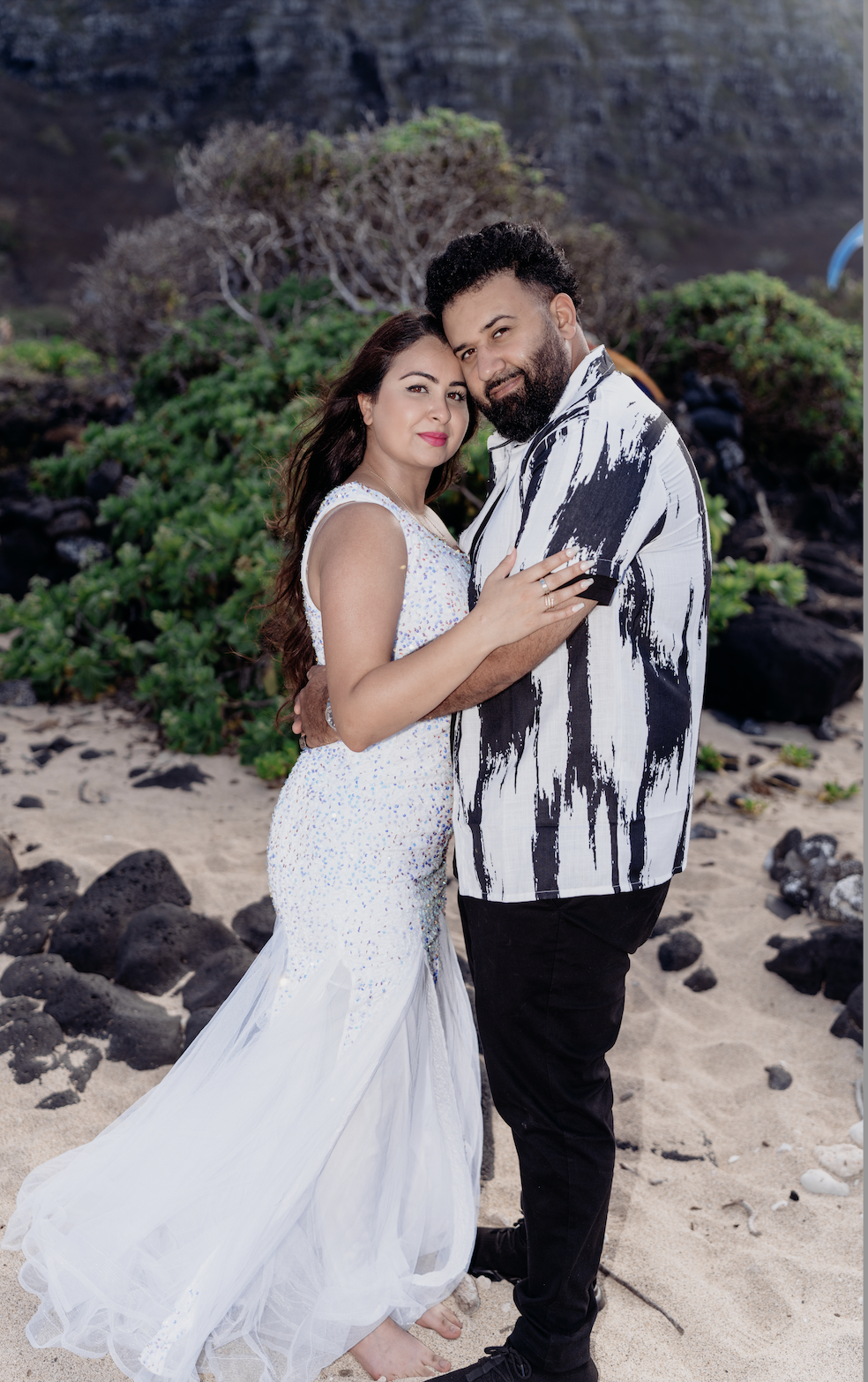 A couple is embracing on a beach with greenery and rocks in the background. The woman wears a white dress with sequins, and the man wears a black and white patterned shirt.