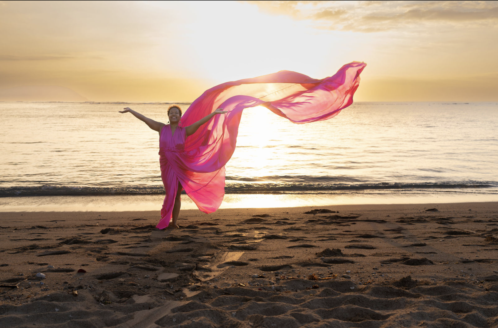 A woman in a pink dress standing on a beach at sunset, holding a flowing pink fabric in the air with arms outstretched, smiling.
