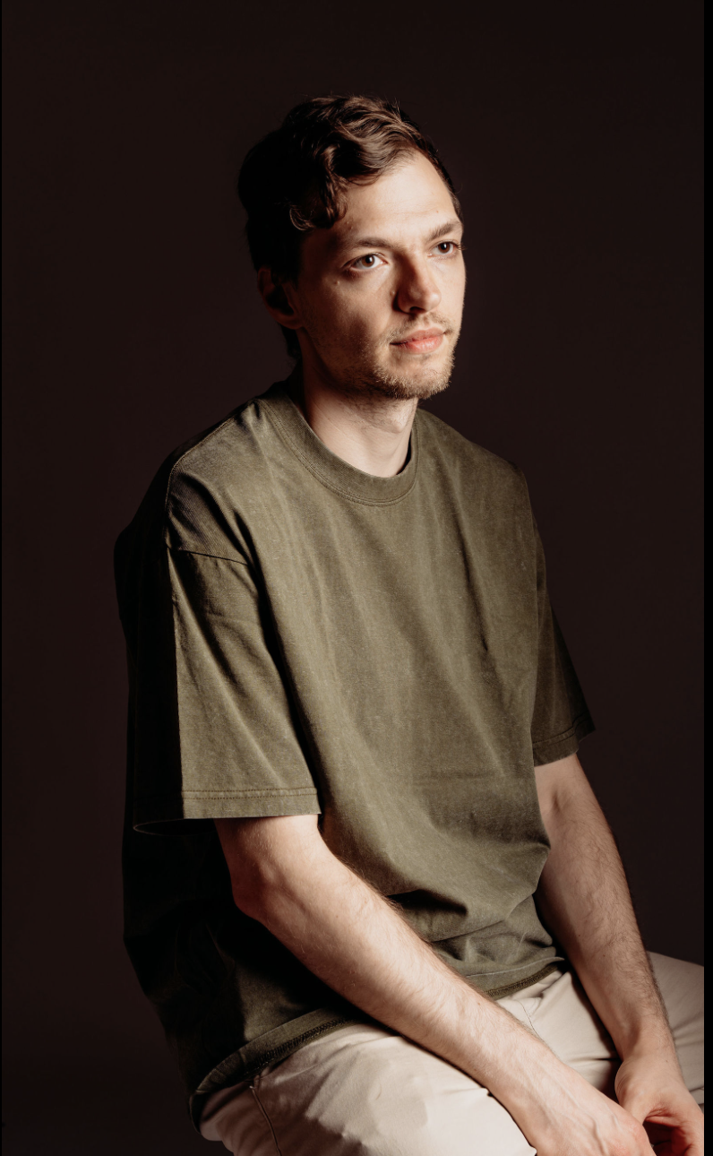 A young man with brown hair and a light beard wearing a loose olive-green t-shirt and beige pants, sitting against a dark backdrop, looking to the side with a thoughtful expression.