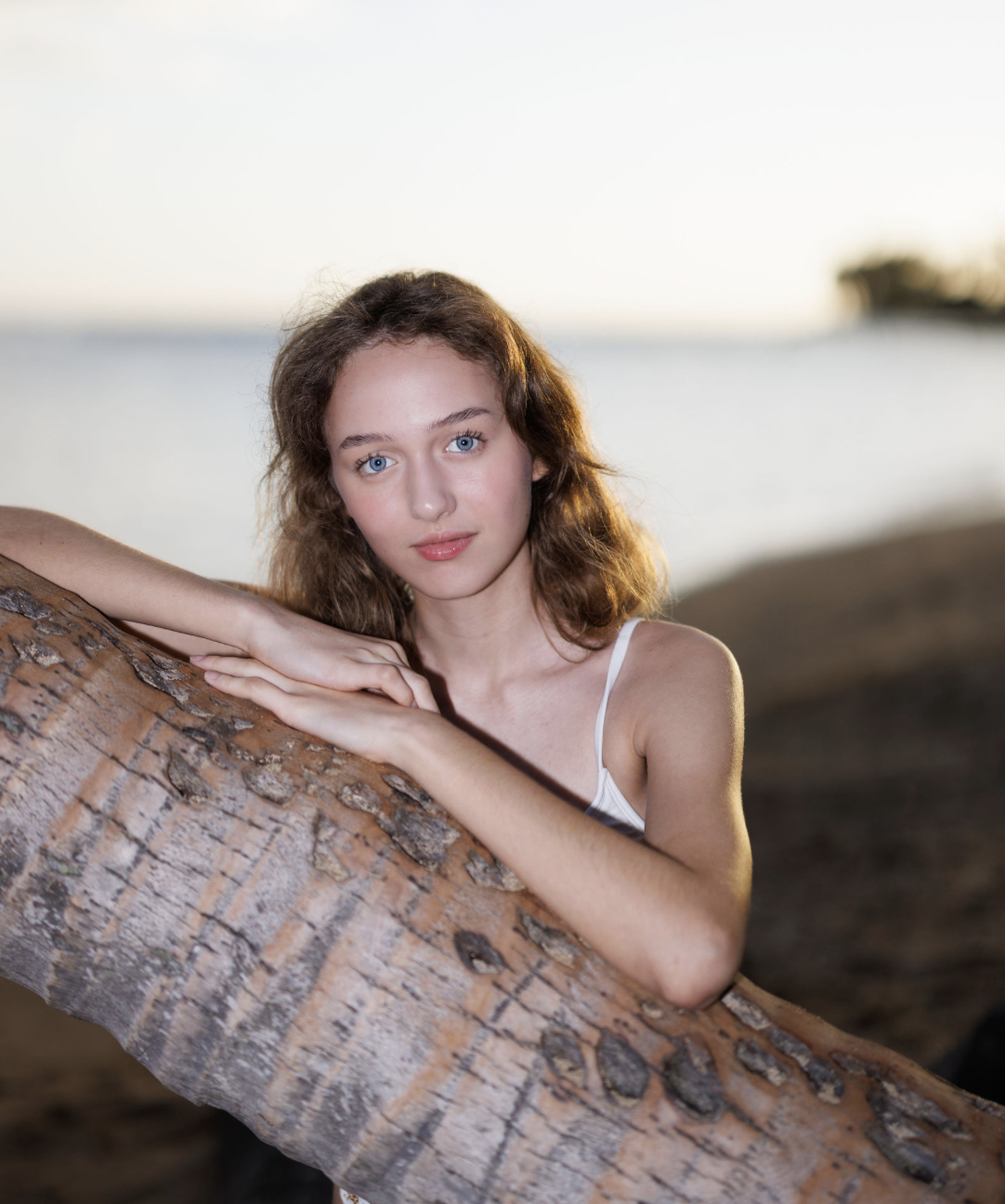 A young woman with curly brown hair and blue eyes leaning on a large driftwood log by the water during sunset.