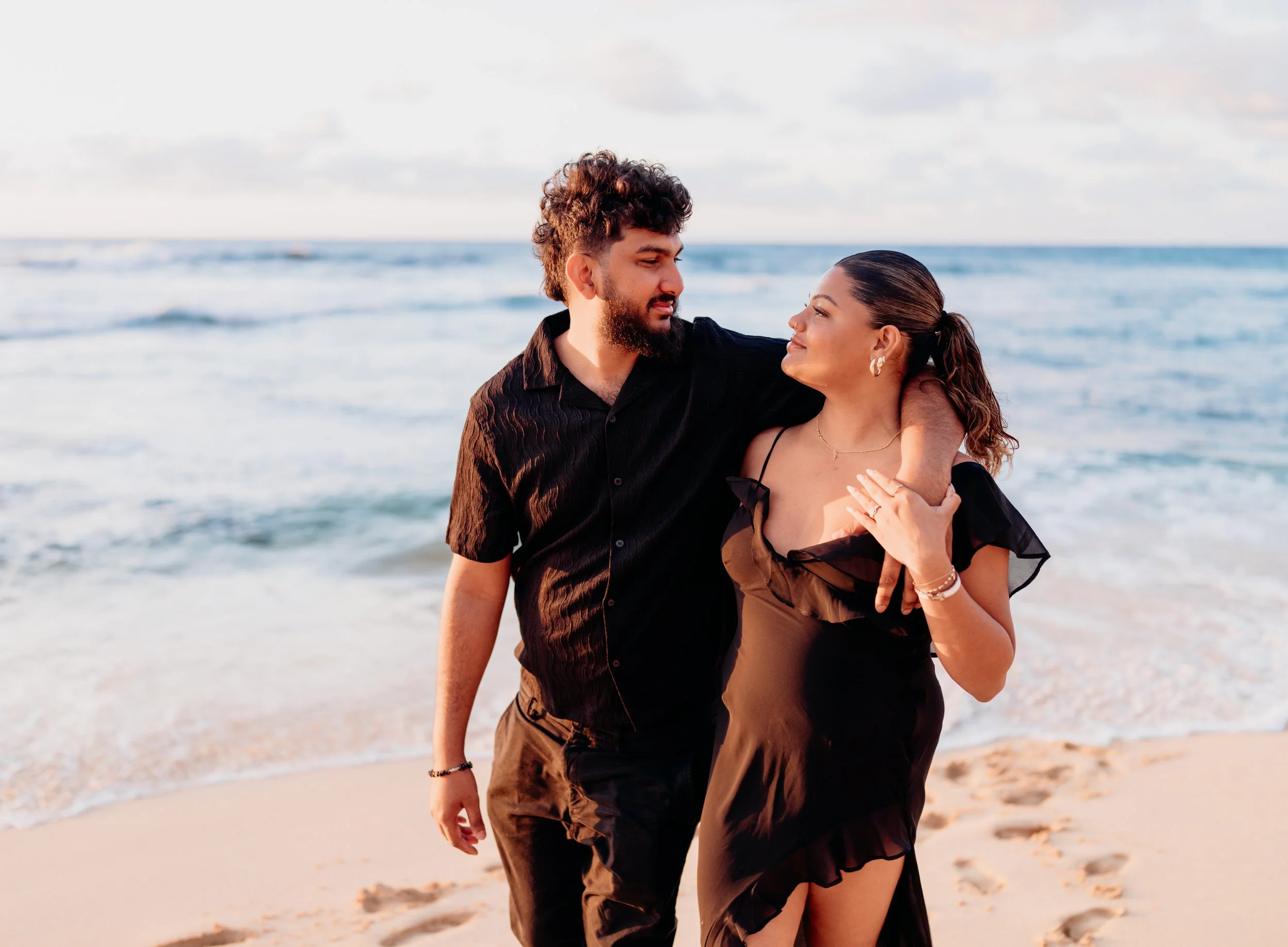 A couple walking on the beach, with the ocean and sky in the background. The man has curly hair and a beard, wearing a black shirt. The woman has long dark hair tied back, wearing a black dress with ruffles. They are looking at each other affectionat