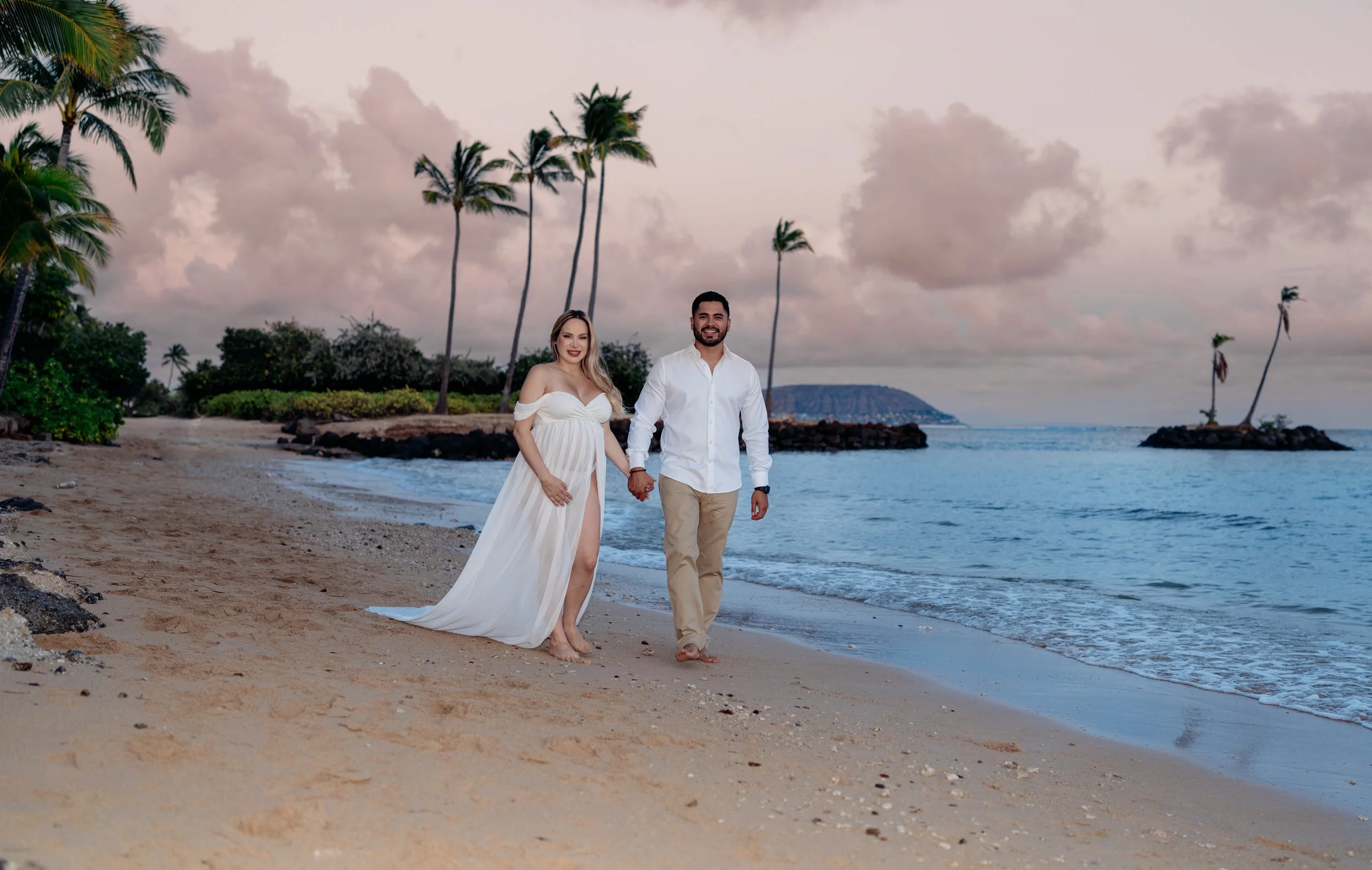 A couple walking hand in hand on a beach at sunset, with palm trees and small islands in the background.
